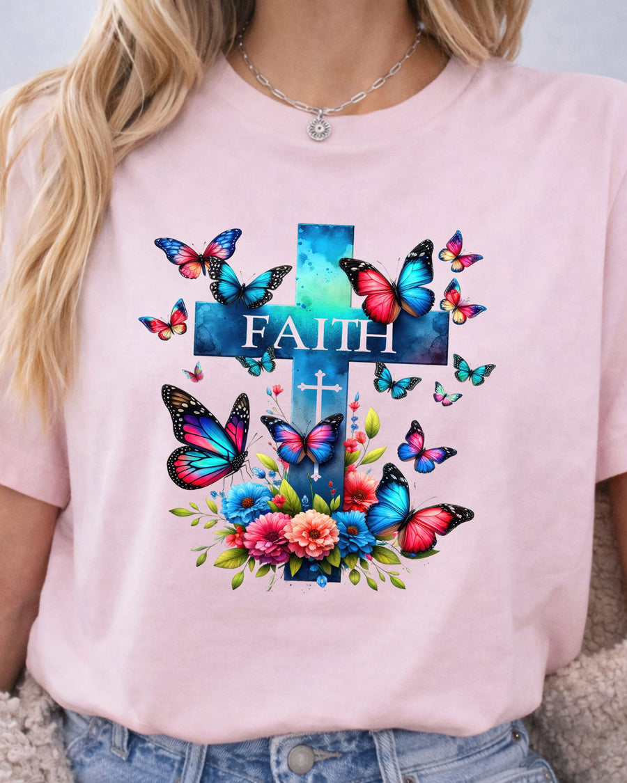 A woman wearing a light pink crewneck t-shirt with a colorful graphic print featuring a blue and teal cross, the word 'FAITH', colorful butterflies, and flowers.