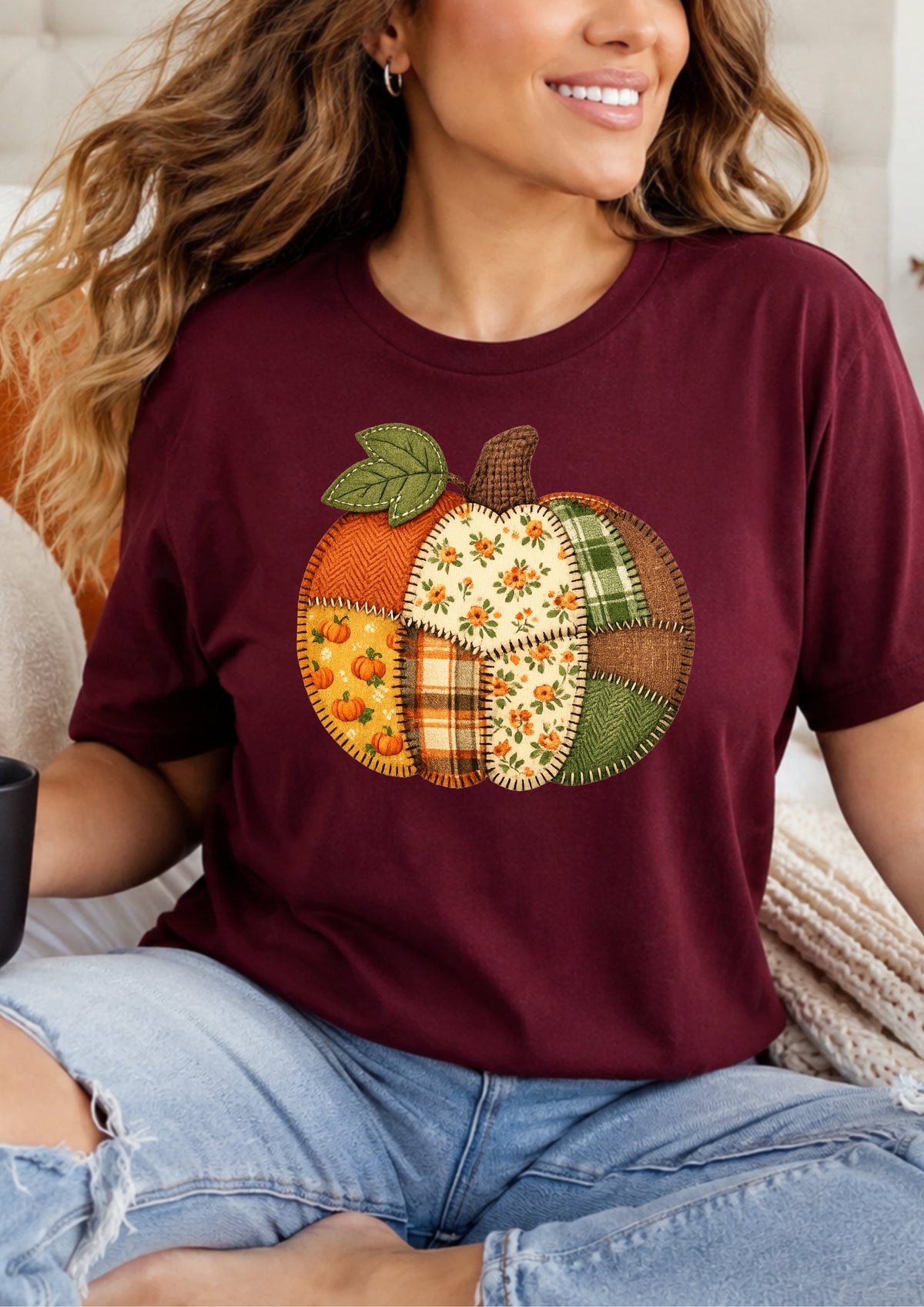 A woman wearing a burgundy short-sleeve t-shirt with a colorful patchwork pumpkin graphic on the front, paired with light-wash distressed denim jeans.
