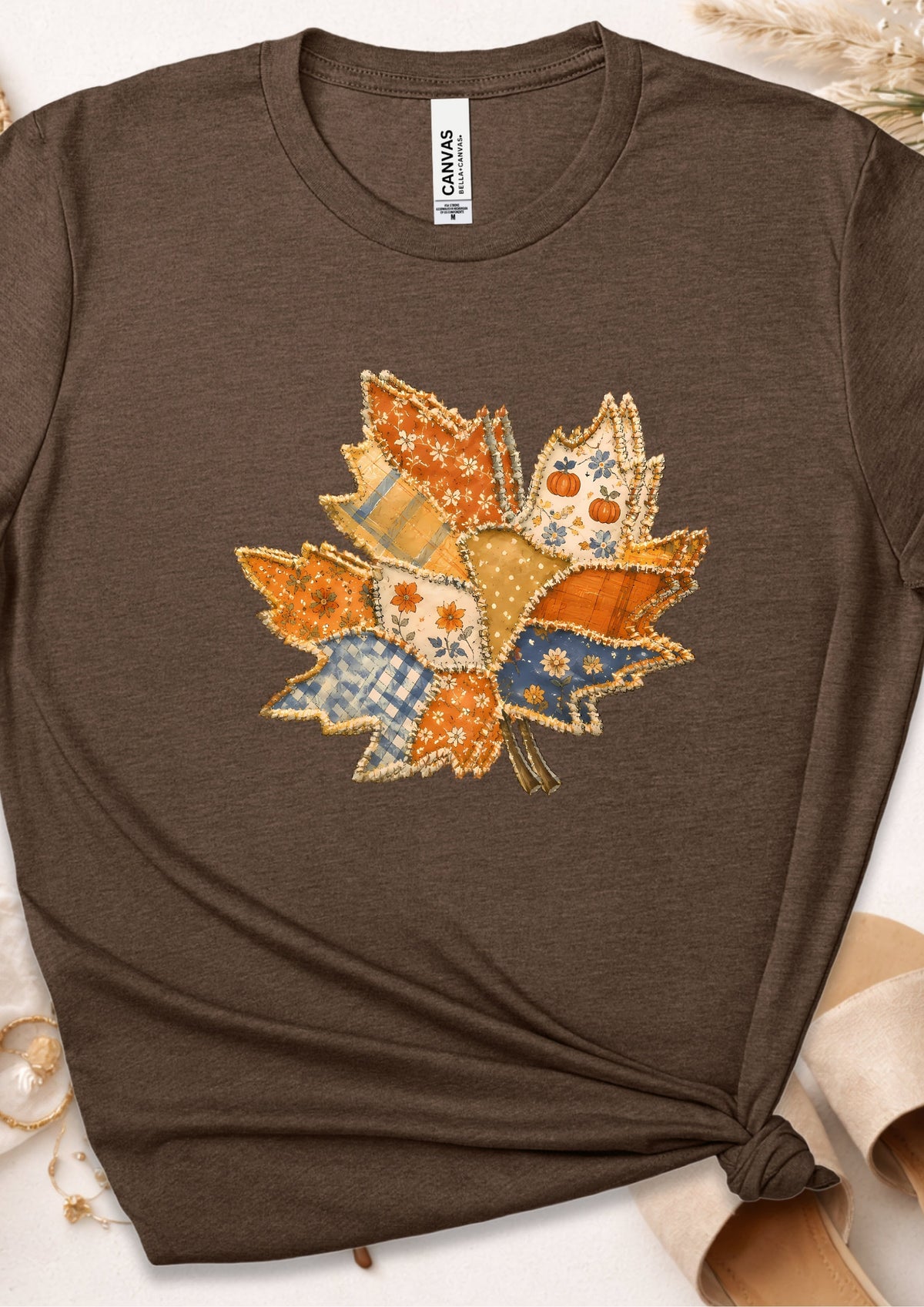 A brown short-sleeve t-shirt with a knotted hem, featuring a colorful autumn leaf patchwork graphic on the chest. The shirt is displayed flat with decorative props in the corners, and a 'CANVAS' neck label is visible.