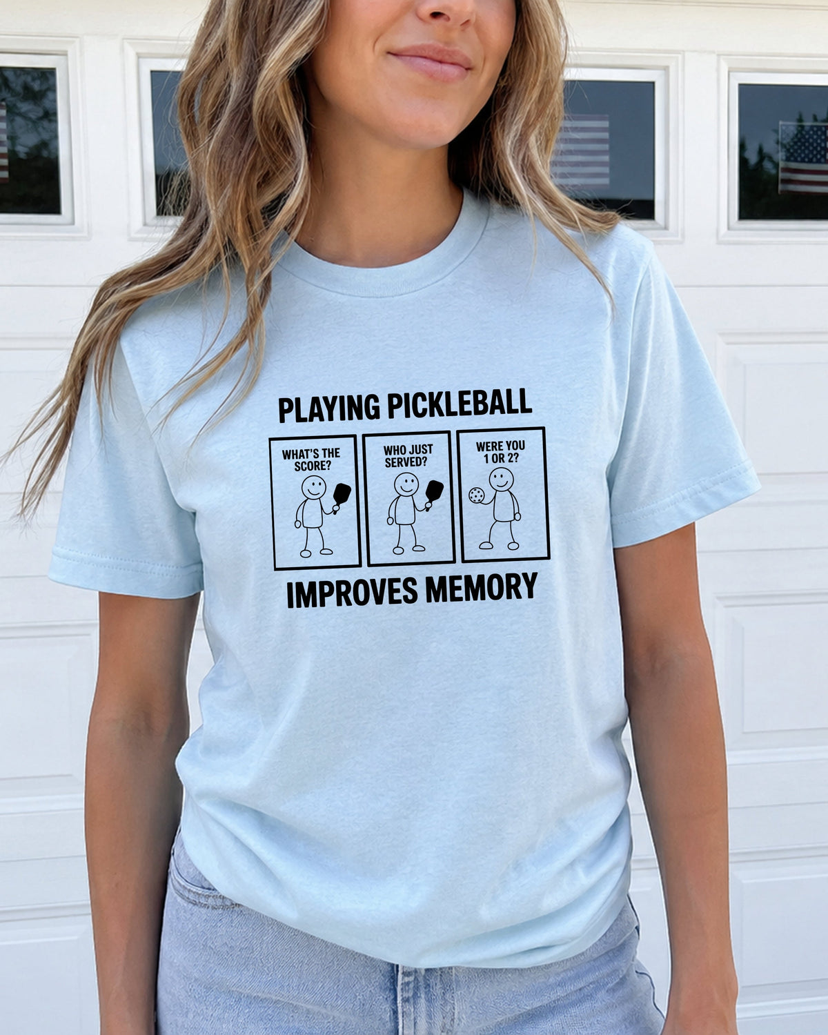 A woman wearing a light blue graphic t-shirt with a black printed design that reads 'PLAYING PICKLEBALL IMPROVES MEMORY' with three cartoon panels, standing in front of a white garage door.