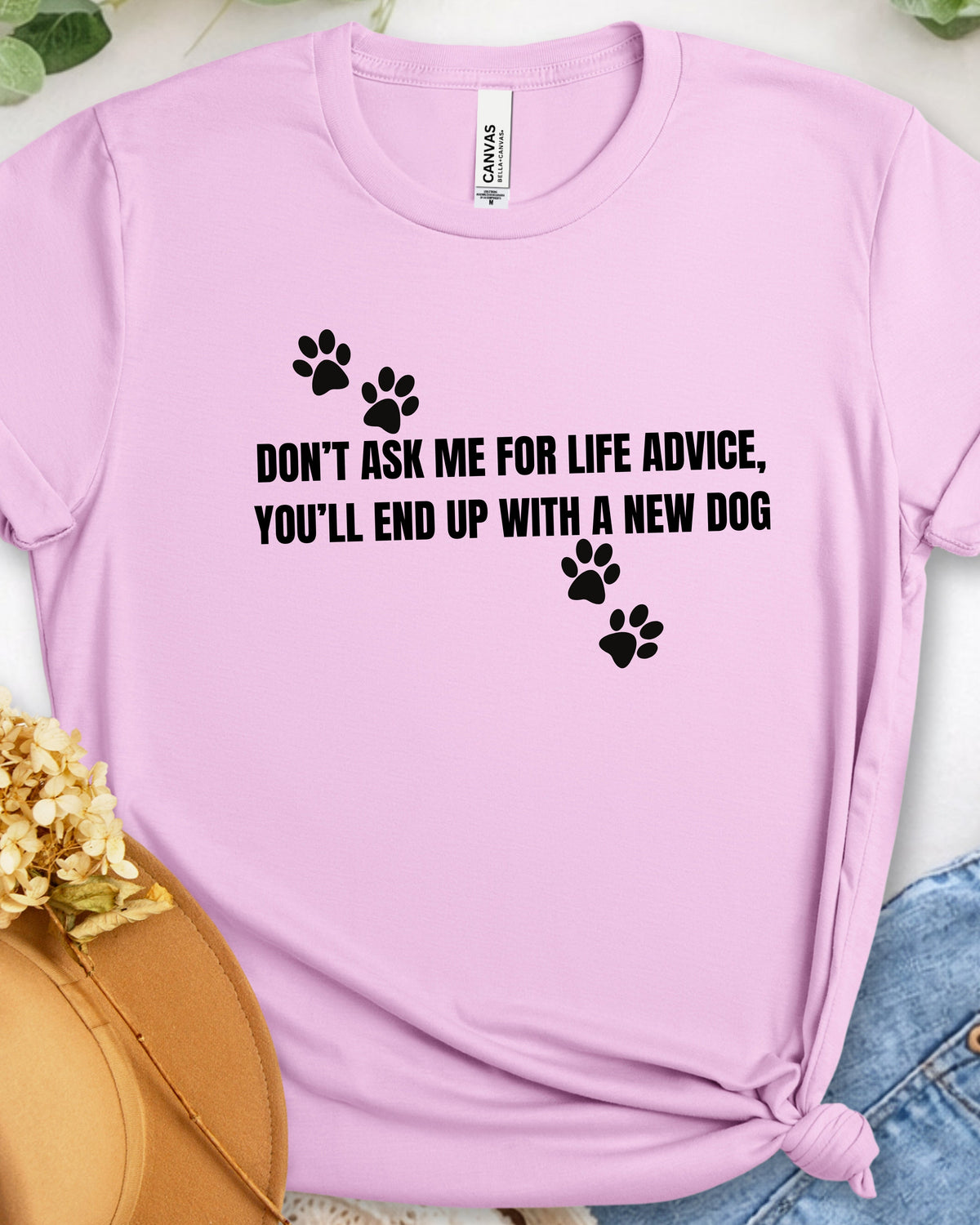 A light pink short-sleeve graphic T-shirt laid flat with a knot at the bottom right. The shirt has black paw prints and the text 'DON'T ASK ME FOR LIFE ADVICE, YOU'LL END UP WITH A NEW DOG' printed on the front.