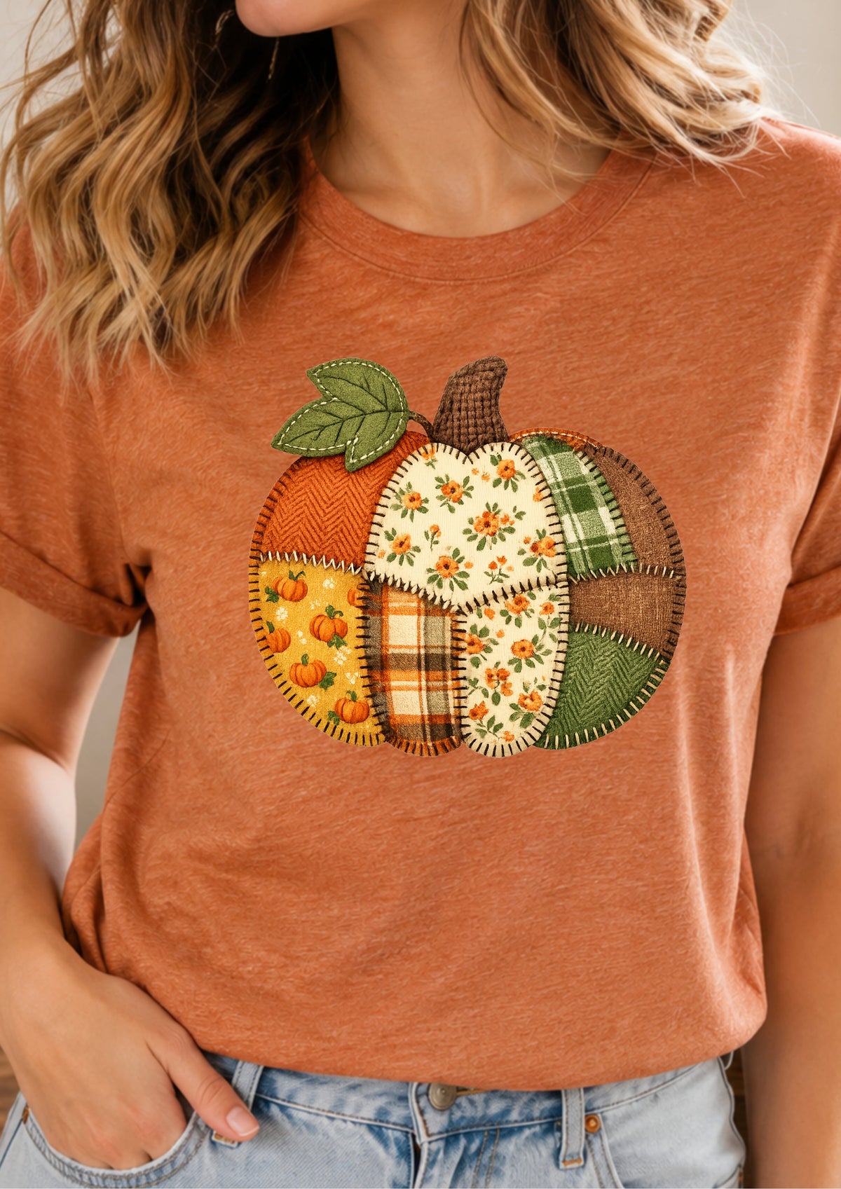 A woman wearing a rust-orange short-sleeve t-shirt with a large, colorful patchwork pumpkin graphic on the front. The pumpkin features various autumn patterns including plaid, floral, and sunflower prints in shades of orange, green, yellow, and brown.