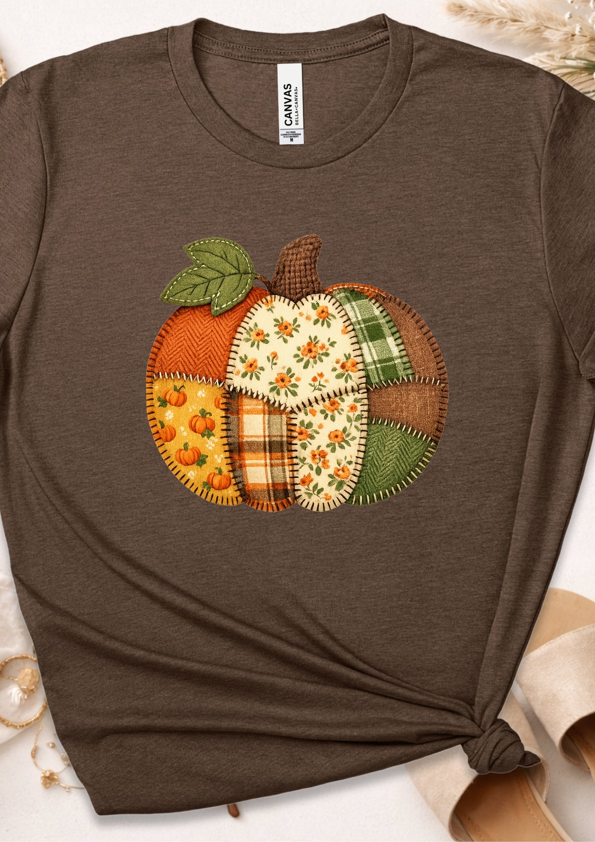 A brown short-sleeve t-shirt featuring a colorful patchwork pumpkin graphic on the chest. The shirt is styled with a knot at the bottom hem and is photographed flat on a light background with subtle autumn-themed props.