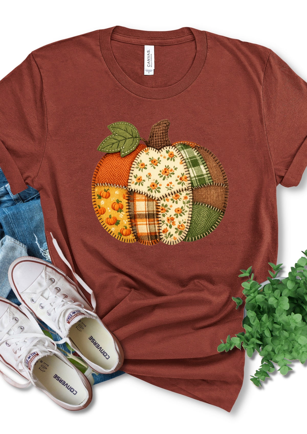 A rust-brown short-sleeve t-shirt featuring a colorful patchwork pumpkin graphic on the chest. The shirt is styled in a flat-lay arrangement with white Converse sneakers, blue jeans, a green plant, and a white neck label that reads 'CANVAS'.