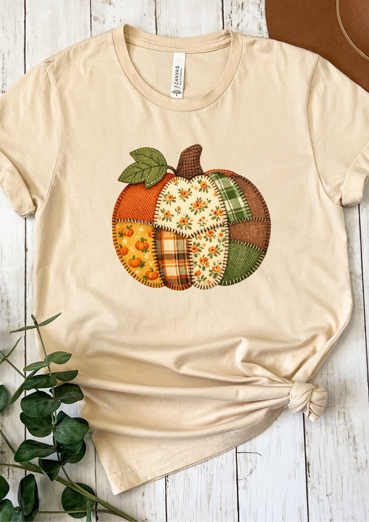 A beige short-sleeve crewneck T-shirt laid flat on a white wood background, featuring a large patchwork pumpkin graphic on the chest with autumn colors and fabric-patterned sections.