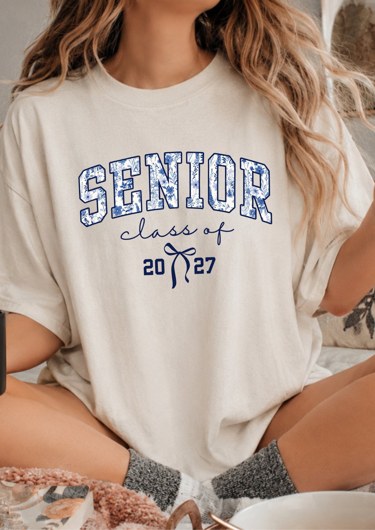 A woman wearing a cream-colored graphic t-shirt with 'SENIOR class of 2027' printed in blue on the front, sitting in a cozy indoor setting.