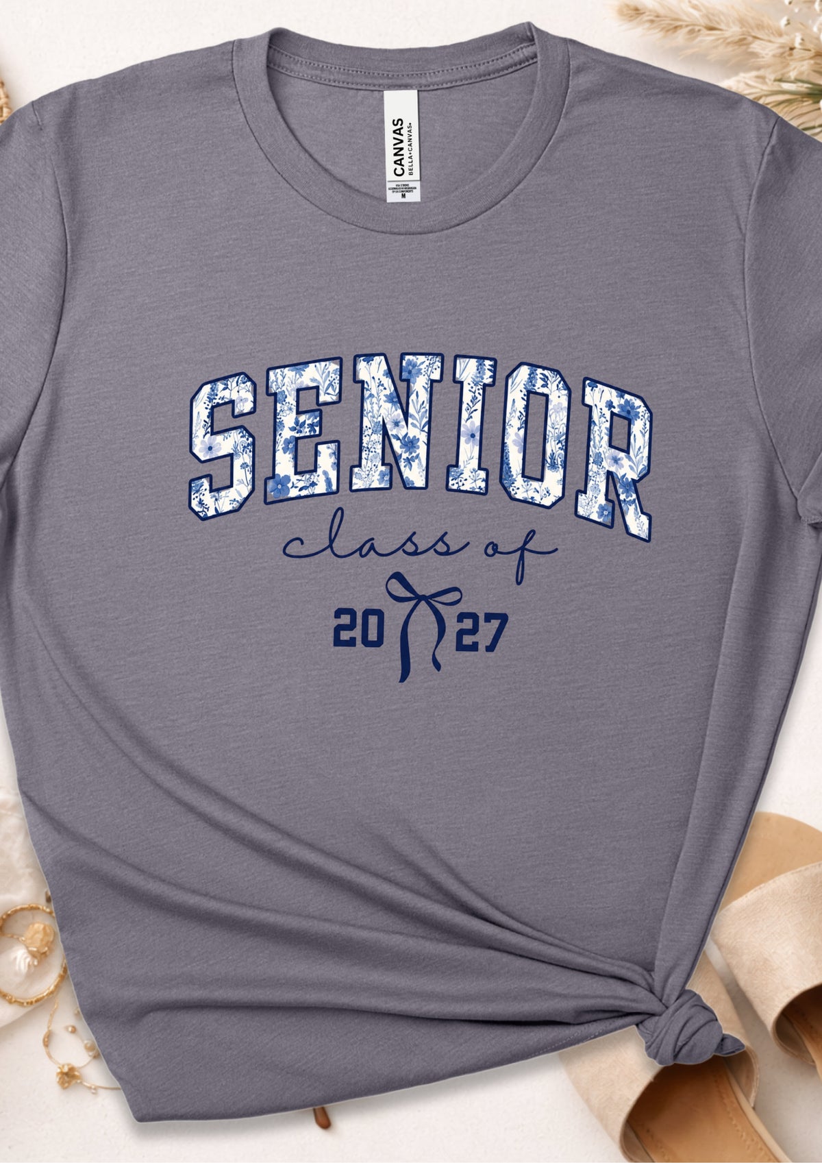 A heather gray crewneck t-shirt with a distressed blue and white 'SENIOR' graphic and 'class of 2027' printed on the front, styled with a knot at the bottom hem and neutral props.