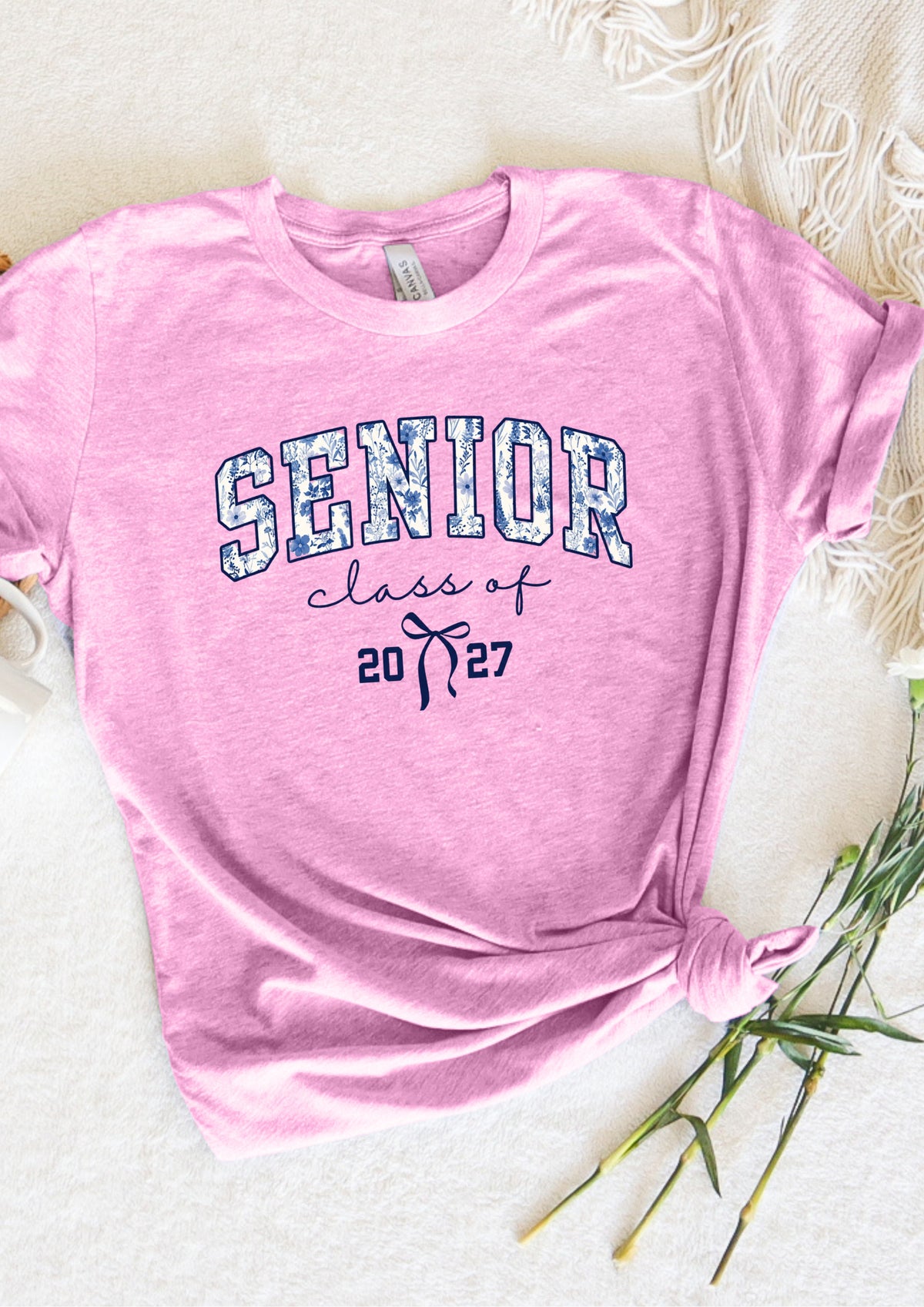 A pink short-sleeve t-shirt with 'SENIOR class of 2027' printed on the front, styled flat on a white textured background with decorative green stems.