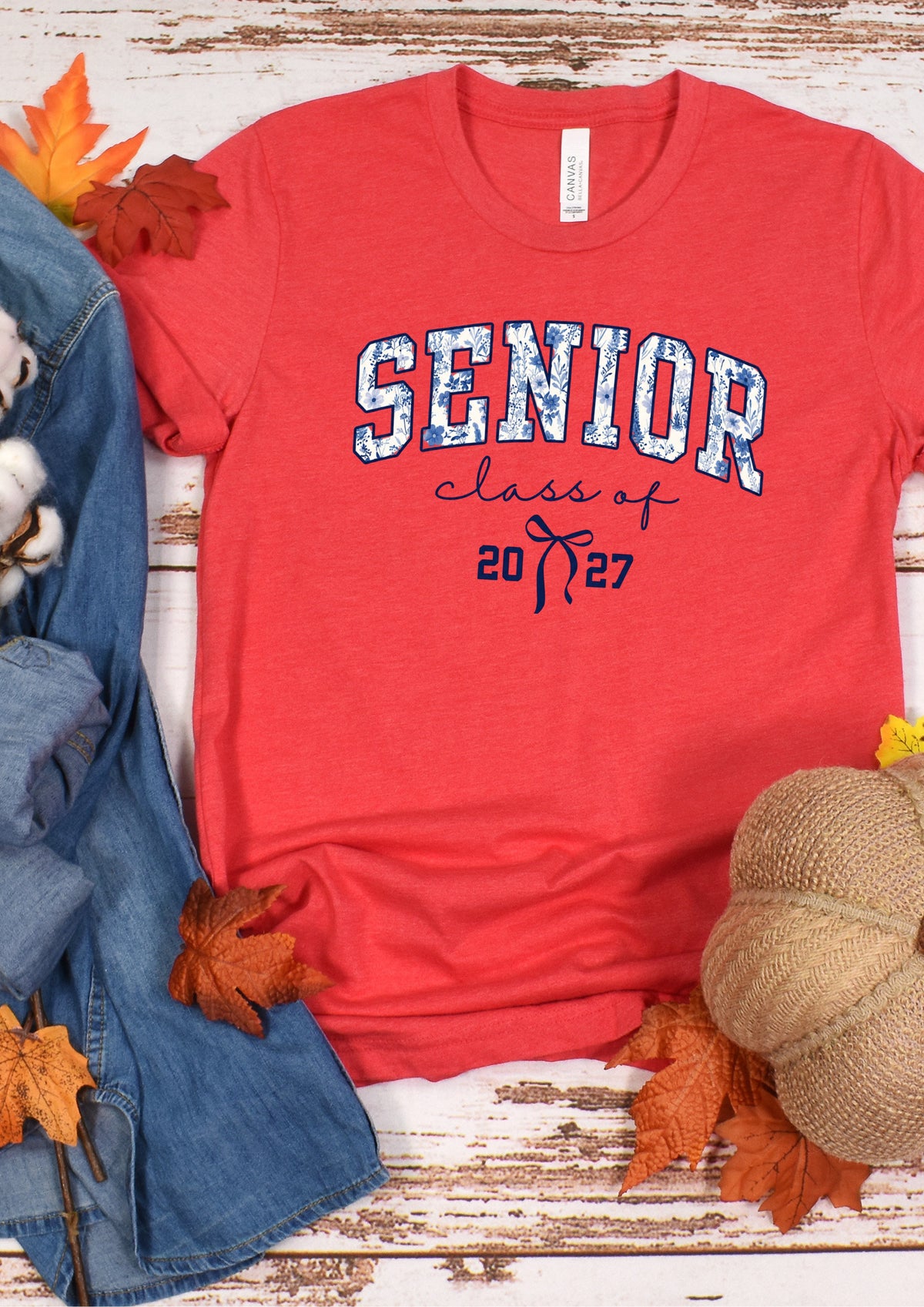 A red short-sleeve graphic T-shirt laid flat on a rustic white wood surface, styled with autumn leaves, a denim jacket, cotton bolls, and a burlap pumpkin. The shirt reads 'SENIOR class of 2027' in white and navy lettering.