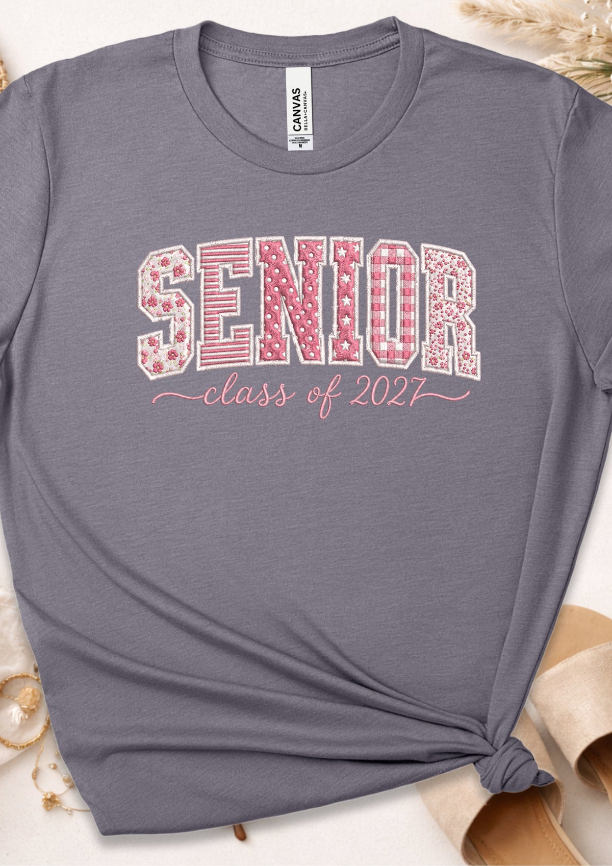 A heather gray t-shirt with a 'SENIOR class of 2027' graphic on the chest. The word 'SENIOR' is filled with a pink and white gingham pattern, and 'class of 2027' is written in a pink cursive font.