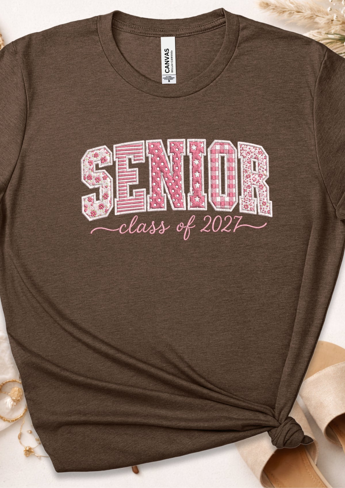 A dark brown heather graphic t-shirt featuring the word 'SENIOR' in pink and white gingham patterned letters, with 'class of 2027' written below in pink script. The t-shirt is styled with a knot at the bottom hem and is shown with a visible Canvas neck label.