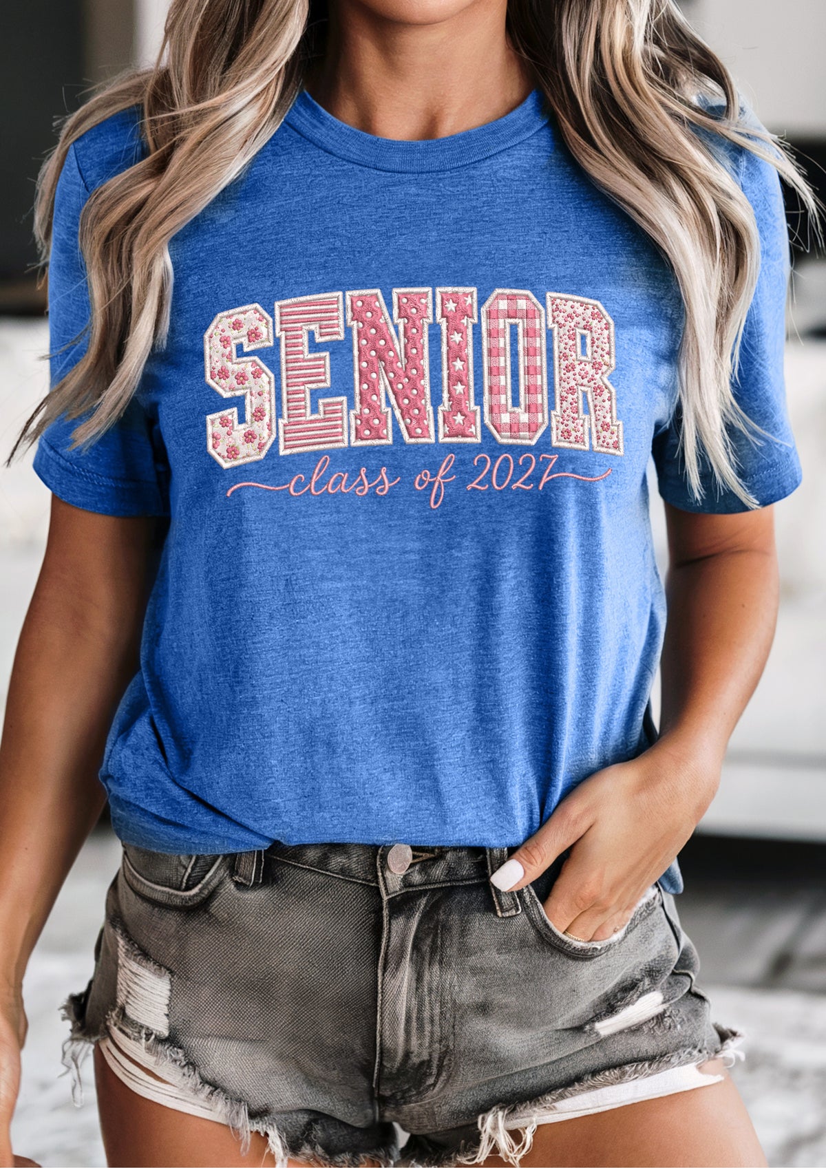 A woman wearing a heather blue graphic t-shirt with 'SENIOR class of 2021' printed in red and white on the front, paired with distressed denim shorts.