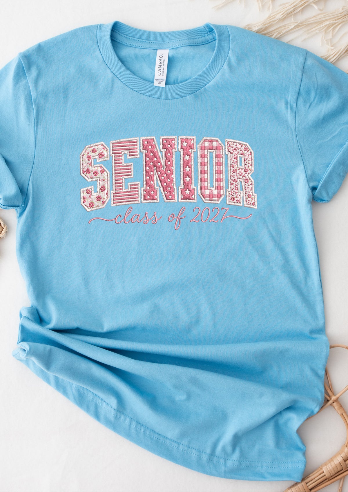 A light blue short-sleeve t-shirt laid flat on a light surface, featuring a graphic that reads 'SENIOR' in pink gingham and floral patterns with 'class of 2021' in pink script underneath.