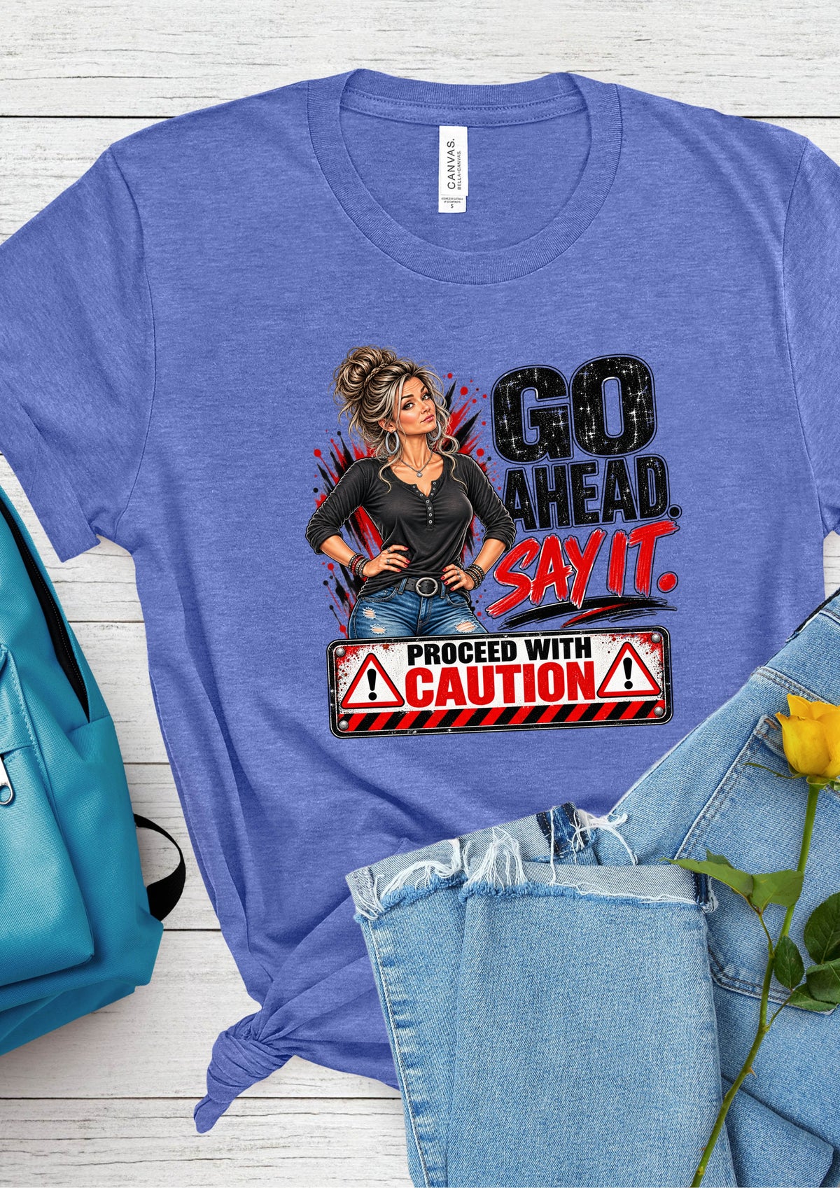 A heathered blue graphic t-shirt laid flat on a white wood background, styled with a teal backpack, light-wash jeans, and a yellow rose. The t-shirt features a graphic of a woman and the text 'GO AHEAD.