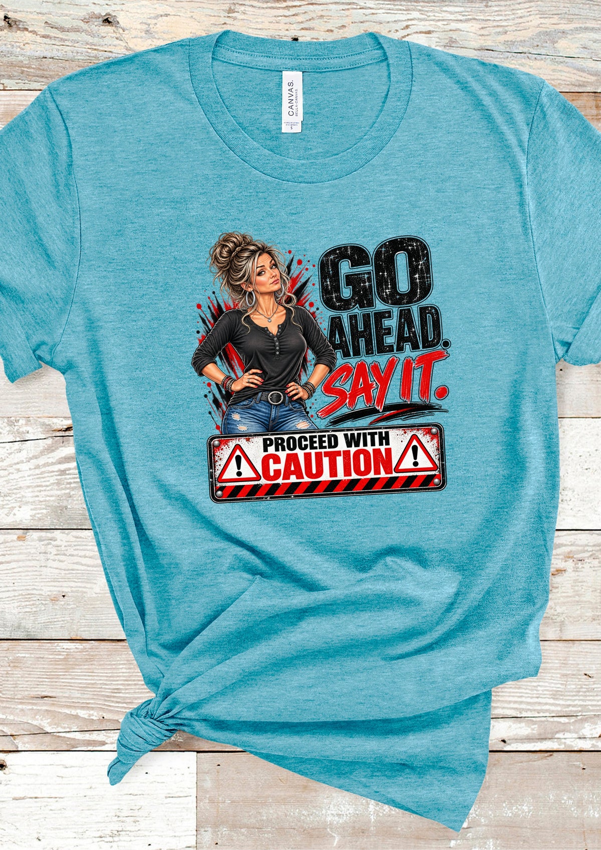 A heathered turquoise blue graphic t-shirt laid flat on a light wood plank background. The shirt features a graphic of a woman with a 'proceed with caution' sign and the text 'GO AHEAD.