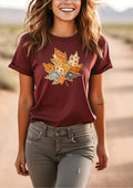 A woman wearing a maroon short-sleeve t-shirt with a colorful patchwork autumn leaf graphic on the front, paired with gray jeans, standing outdoors on a dirt path.
