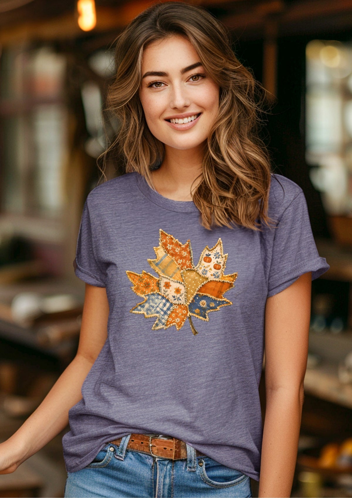 A woman wearing a heathered periwinkle purple t-shirt with a colorful autumn leaf patchwork graphic on the front, paired with blue jeans and a brown belt, in a warm, softly blurred indoor setting.
