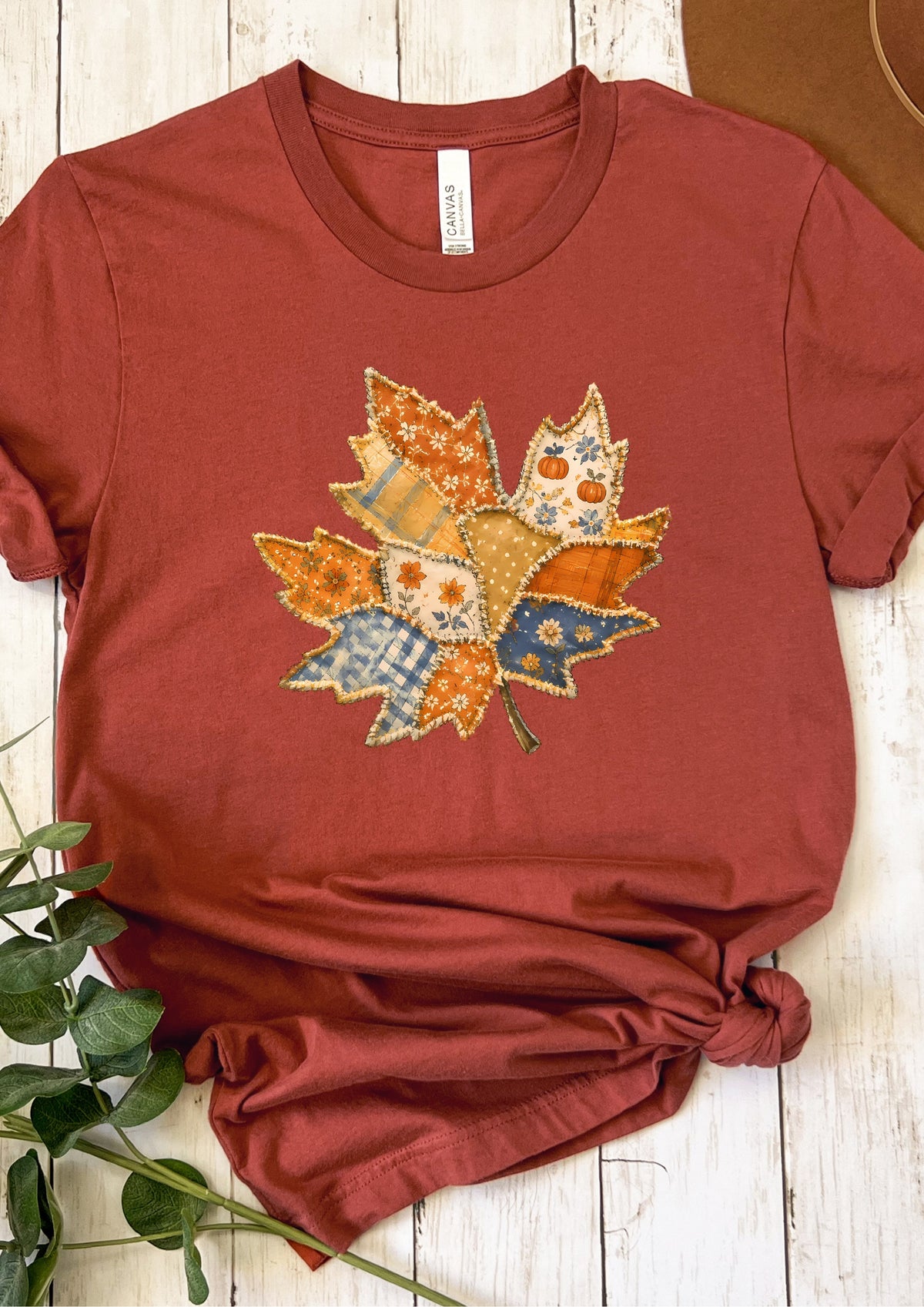 A rust-colored short-sleeve t-shirt with a central autumn leaf applique featuring two owls, laid flat on a white wooden surface with green leaves in the corner.