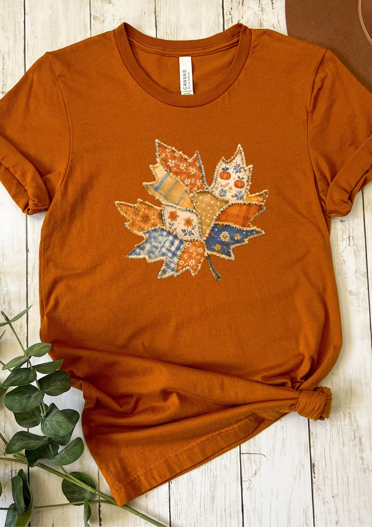 An orange short-sleeve t-shirt with a colorful autumn leaf patchwork graphic on the chest, styled on a white wooden background with green leaves in the corner.