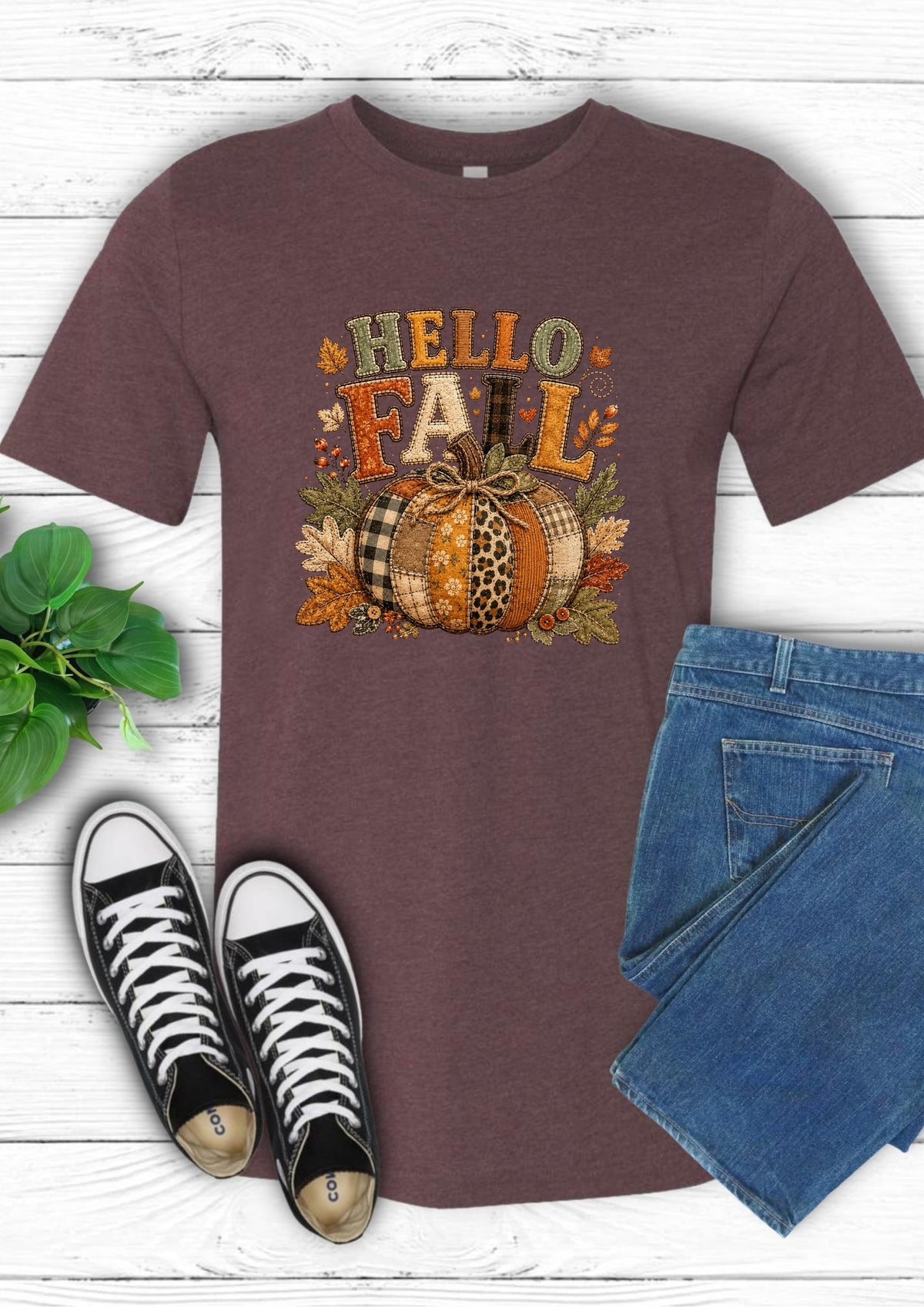 A heathered dark brown short-sleeve t-shirt with a 'HELLO FALL' graphic featuring a pumpkin, leaves, and autumn colors, styled in a flat lay with blue jeans, black sneakers, and a green plant on a white wood background.