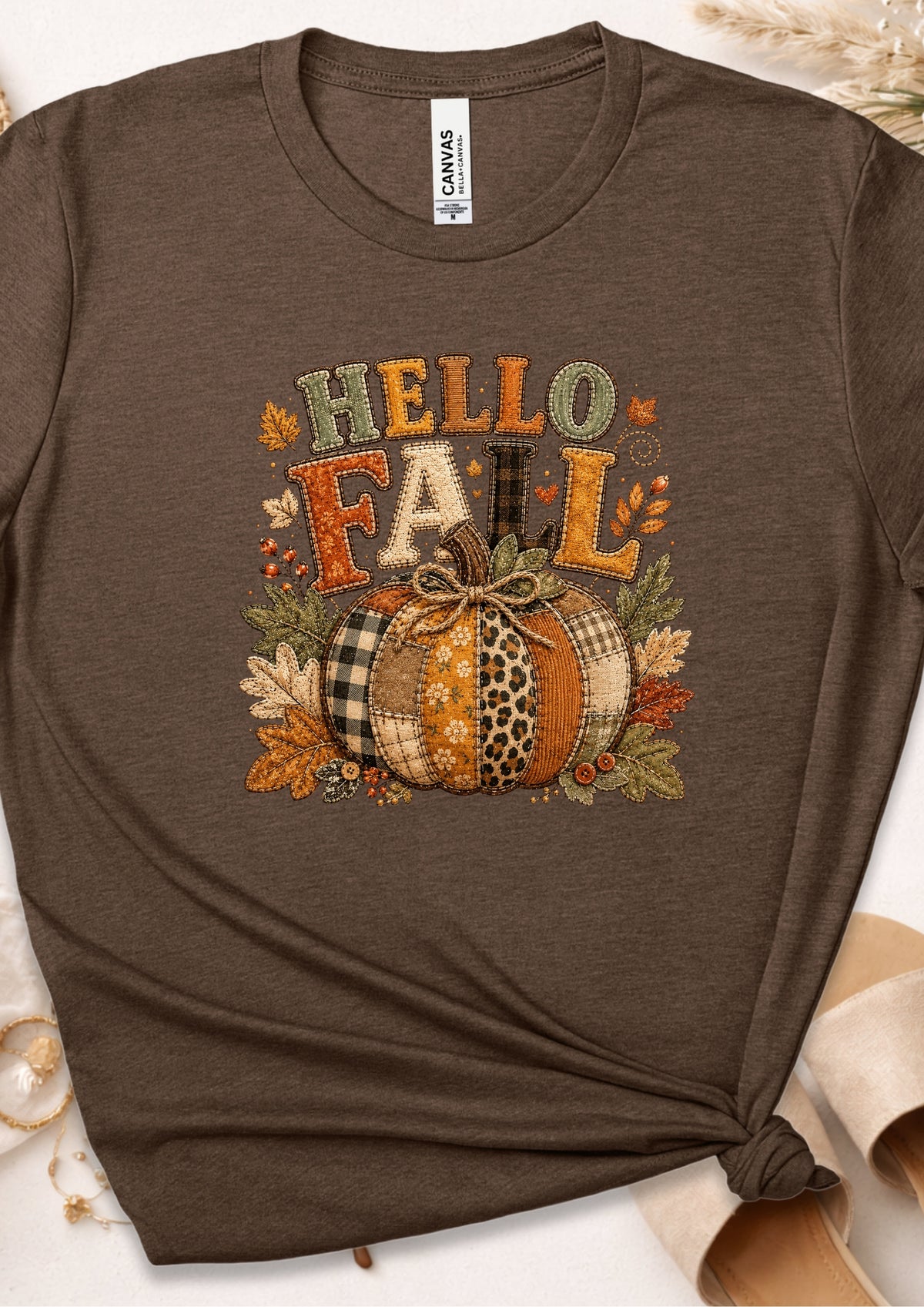 A dark brown heather graphic t-shirt featuring a 'HELLO FALL' autumn design with a patterned pumpkin, leaves, and plaid accents, styled flat with a knot at the bottom right.