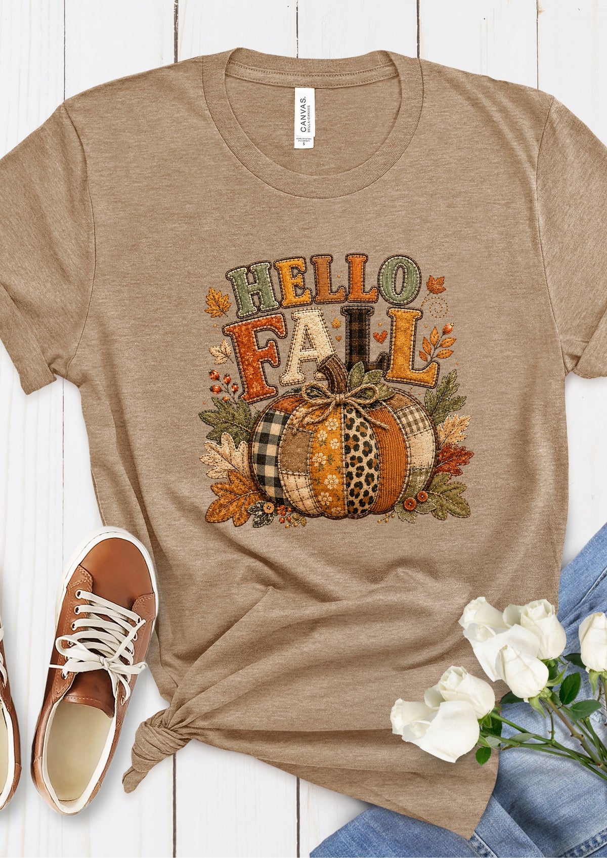 A taupe heather short-sleeve t-shirt with a 'HELLO FALL' autumn graphic featuring a plaid and leopard-print pumpkin, styled in a flat-lay with brown sneakers, white roses, and blue jeans on a white wood background.