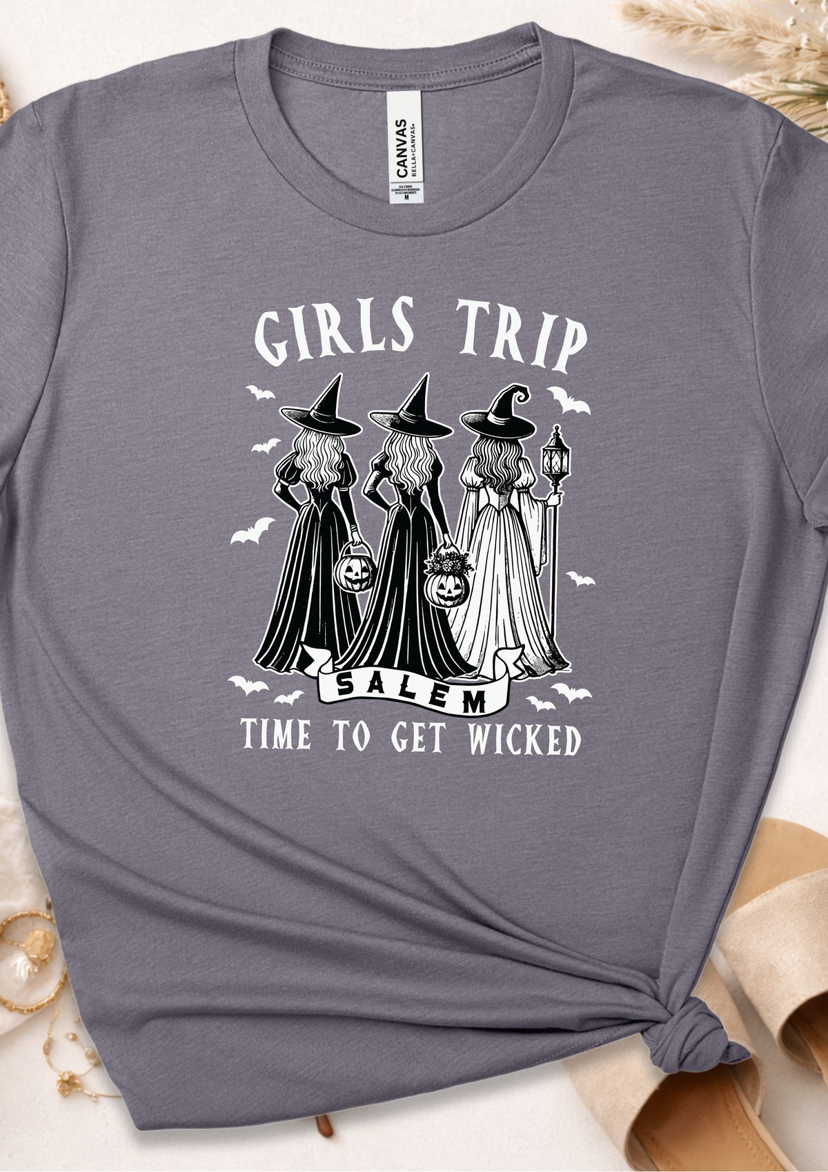 A heather gray graphic t-shirt featuring three witch illustrations with the text 'GIRLS TRIP TO SALEM' and 'TIME TO GET WICKED'. The t-shirt is styled flat with a knot at the bottom right, surrounded by neutral-toned props.