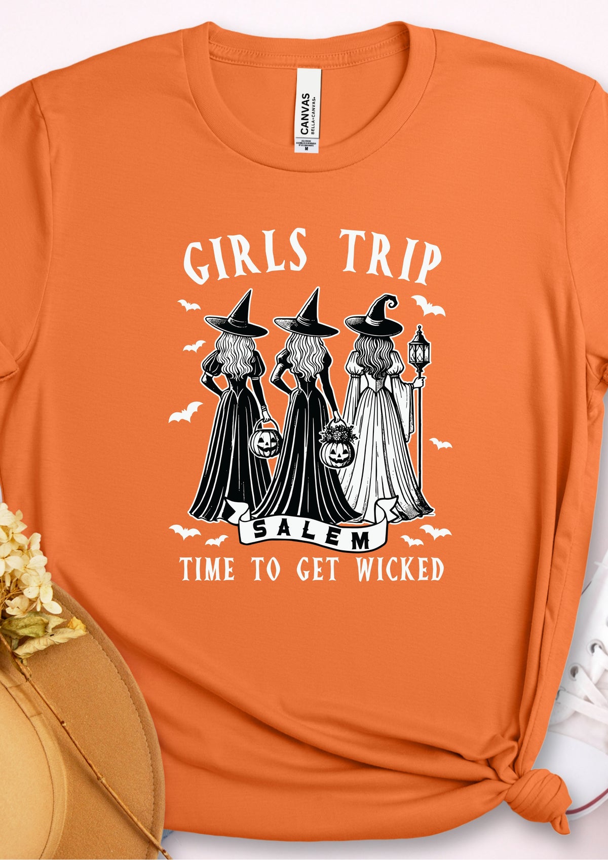 An orange short-sleeve t-shirt with a Halloween graphic featuring three witches in black and white cloaks with the text 'GIRLS TRIP SALEM TIME TO GET WICKED'. The shirt is styled flat with a hat and shoe visible at the edges.