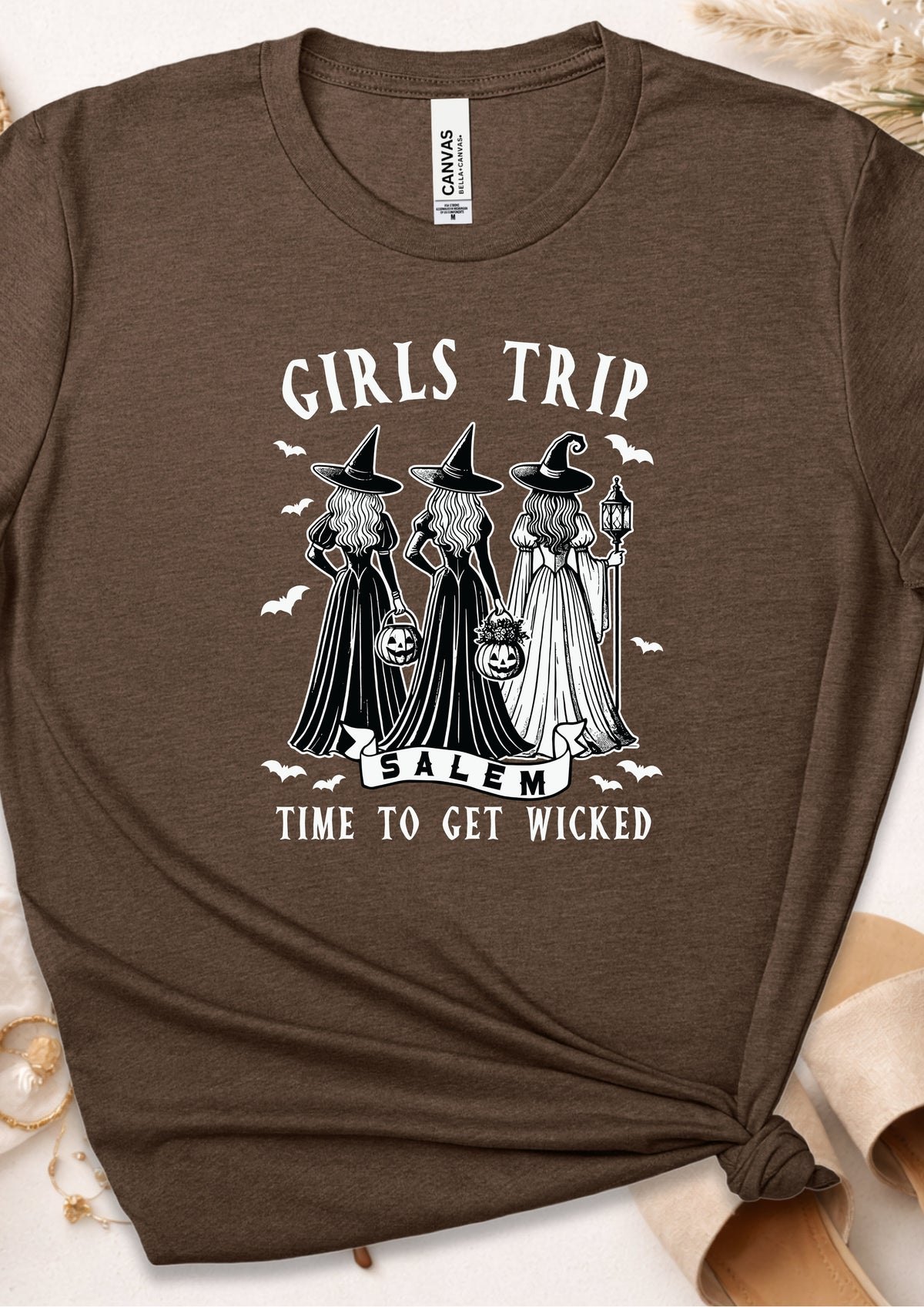 A brown short-sleeve graphic T-shirt laid flat with a knot at the bottom right. The shirt features a white printed design of three witches in hats and gowns with the text 'GIRLS TRIP SALEM' and 'TIME TO GET WICKED.