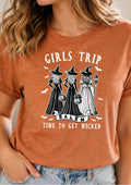 A woman wearing a heathered rust-orange graphic t-shirt with a black and white illustration of three witches in Salem holding pumpkins and a lantern, with the text 'GIRLS TRIP' and 'TIME TO GET WICKED'.