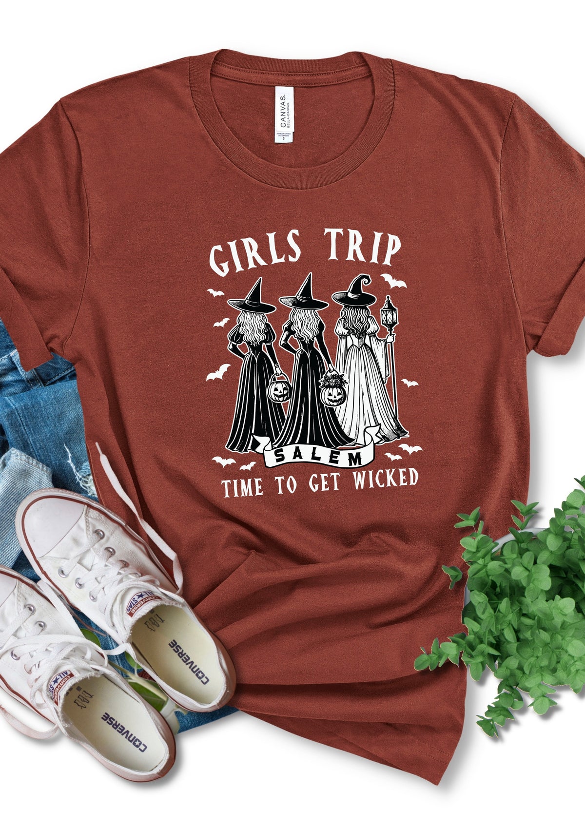 A rust-brown short-sleeve graphic t-shirt laid flat with jeans, white sneakers, and green leaves around it. The shirt features a black and white illustration of three witches with hats and bags, with the text 'GIRLS TRIP SALEM' and 'TIME TO GET WICKED'.