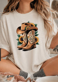 A woman wearing a cream-colored t-shirt with a graphic print of brown cowboy boots, a brown cowboy hat, and yellow sunflowers. The setting appears to be a cozy indoor environment with a bed or couch in the background.