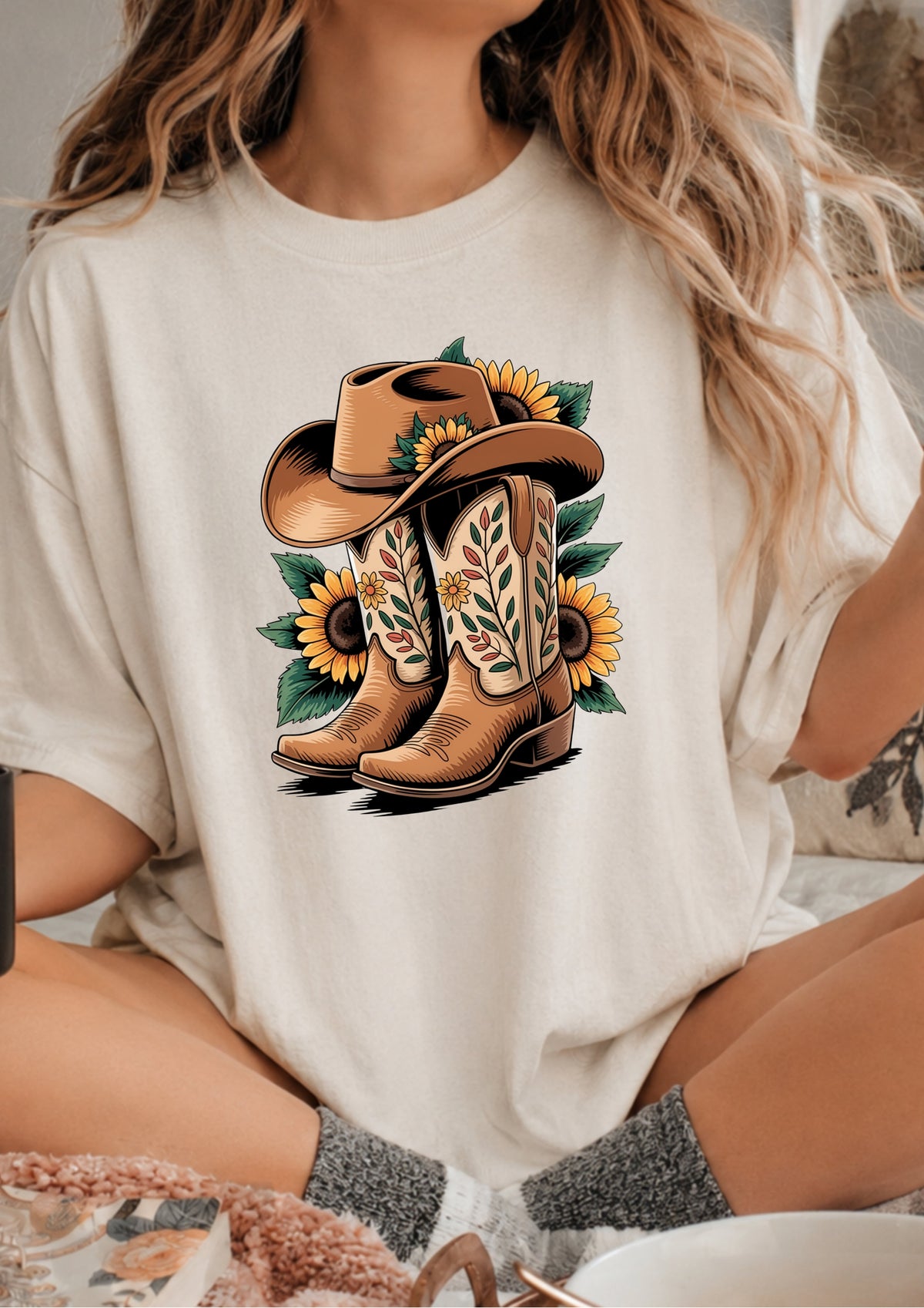 A woman wearing a cream-colored t-shirt with a graphic print of brown cowboy boots, a brown cowboy hat, and yellow sunflowers. The setting appears to be a cozy indoor environment with a bed or couch in the background.