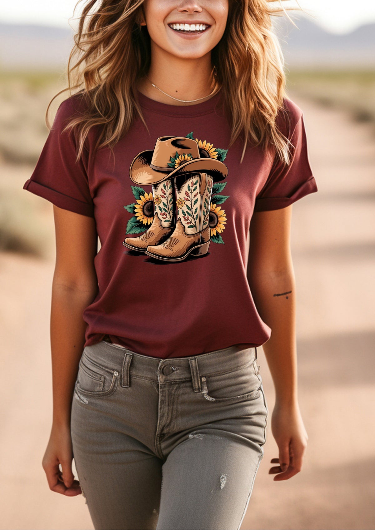 A woman wearing a maroon short-sleeve graphic t-shirt with a western print of cowboy boots, a hat, and sunflowers, paired with gray jeans, set against a blurred desert background.