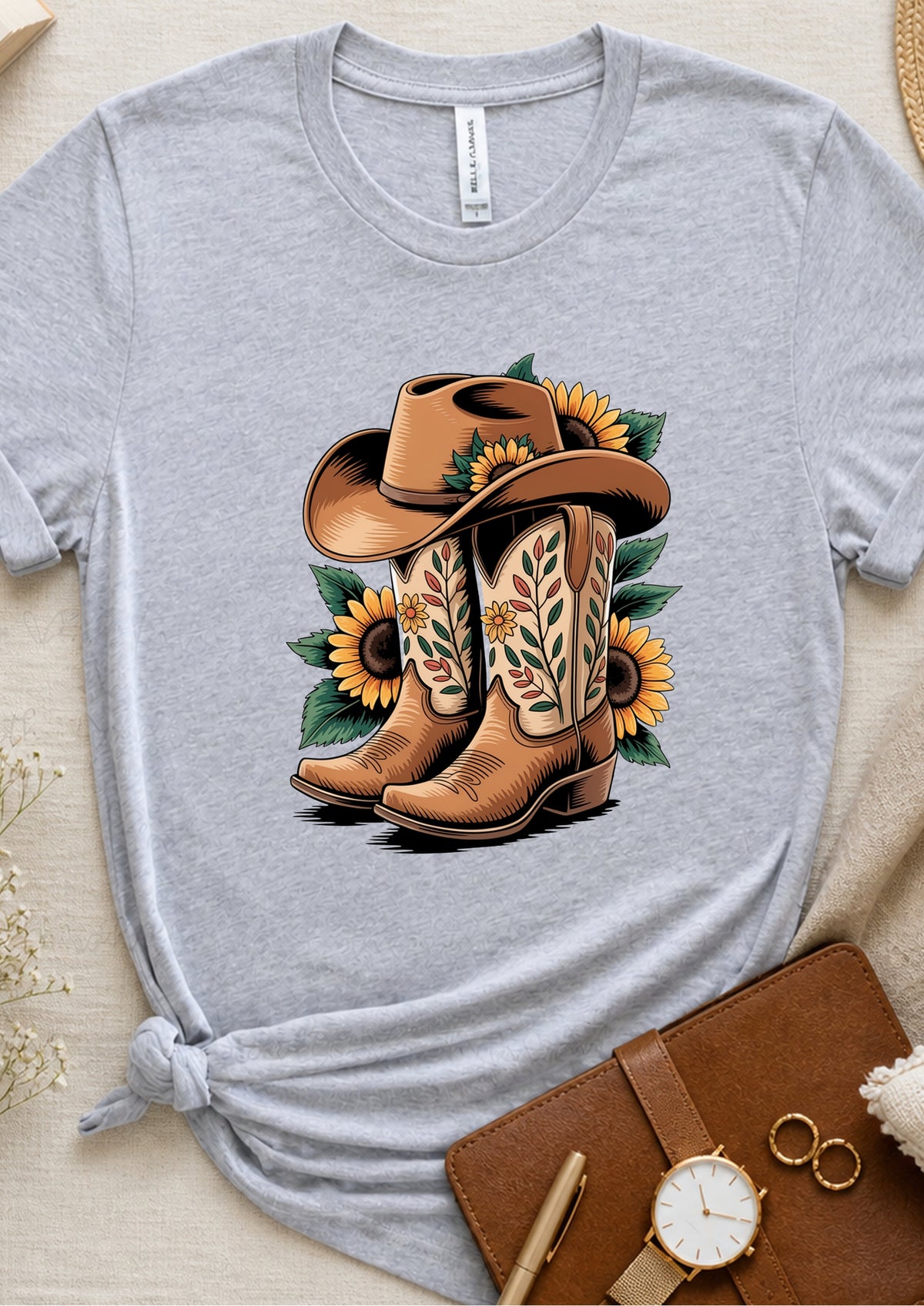 A heather gray short-sleeve t-shirt with a western-themed graphic of brown cowboy boots, a cowboy hat, and yellow sunflowers, styled flat-lay on a beige fabric background with a brown notebook, watch, and rings.