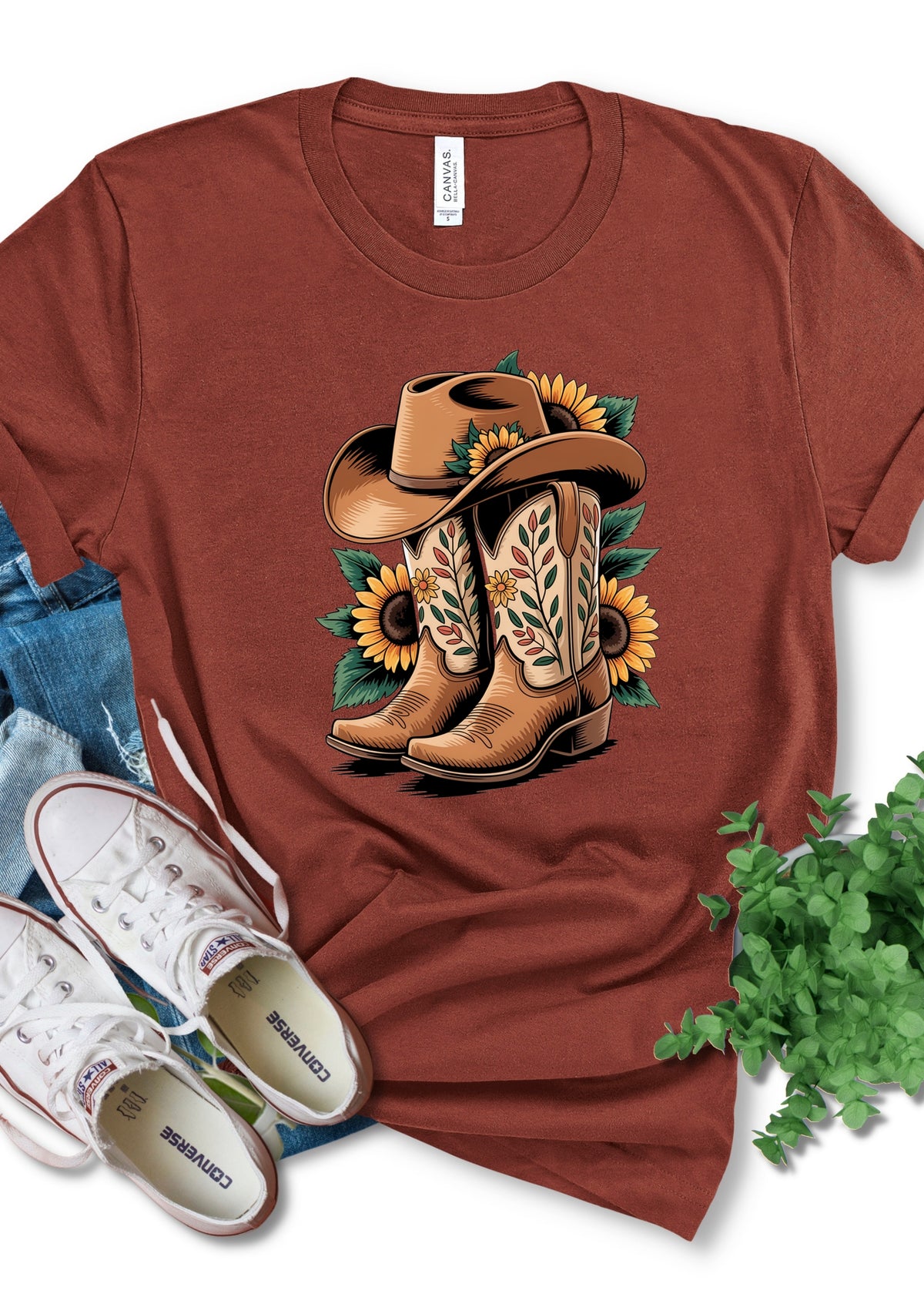 A rust-brown short-sleeve t-shirt with a graphic of cowboy boots and a cowboy hat decorated with sunflowers, styled in a flat lay with jeans, white sneakers, and green leaves.