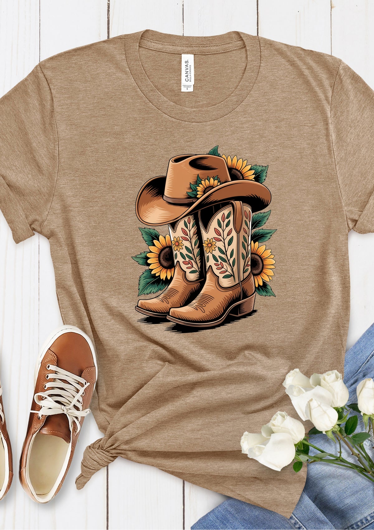 A heather tan short-sleeve t-shirt with a western graphic of a cowboy hat, boots, and sunflowers, styled in a flat-lay with brown sneakers, white roses, and blue jeans on a white wood background.