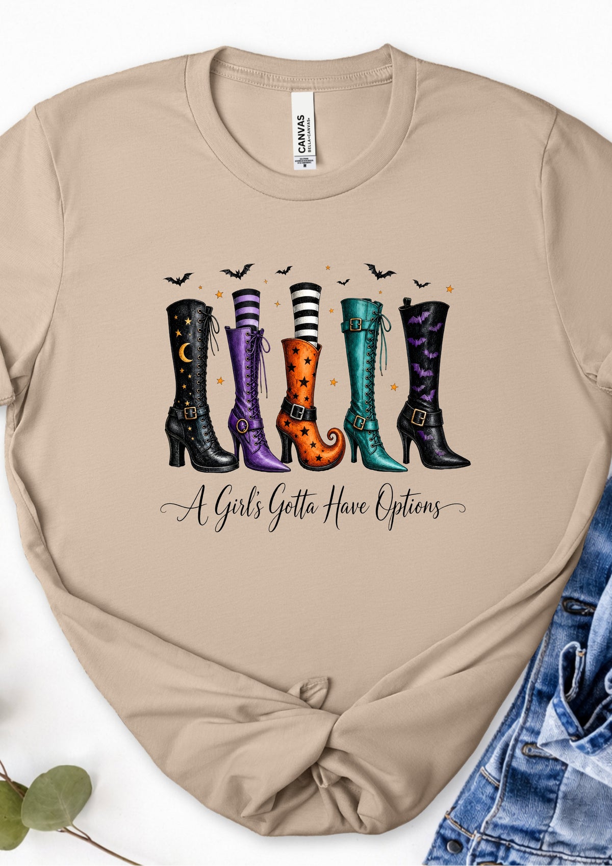 A beige crewneck t-shirt featuring a graphic of four colorful witch boots with the text 'A Girl's Gotta Have Options' underneath. The shirt is styled flat with a denim jacket on the right and a green leaf on the bottom left.