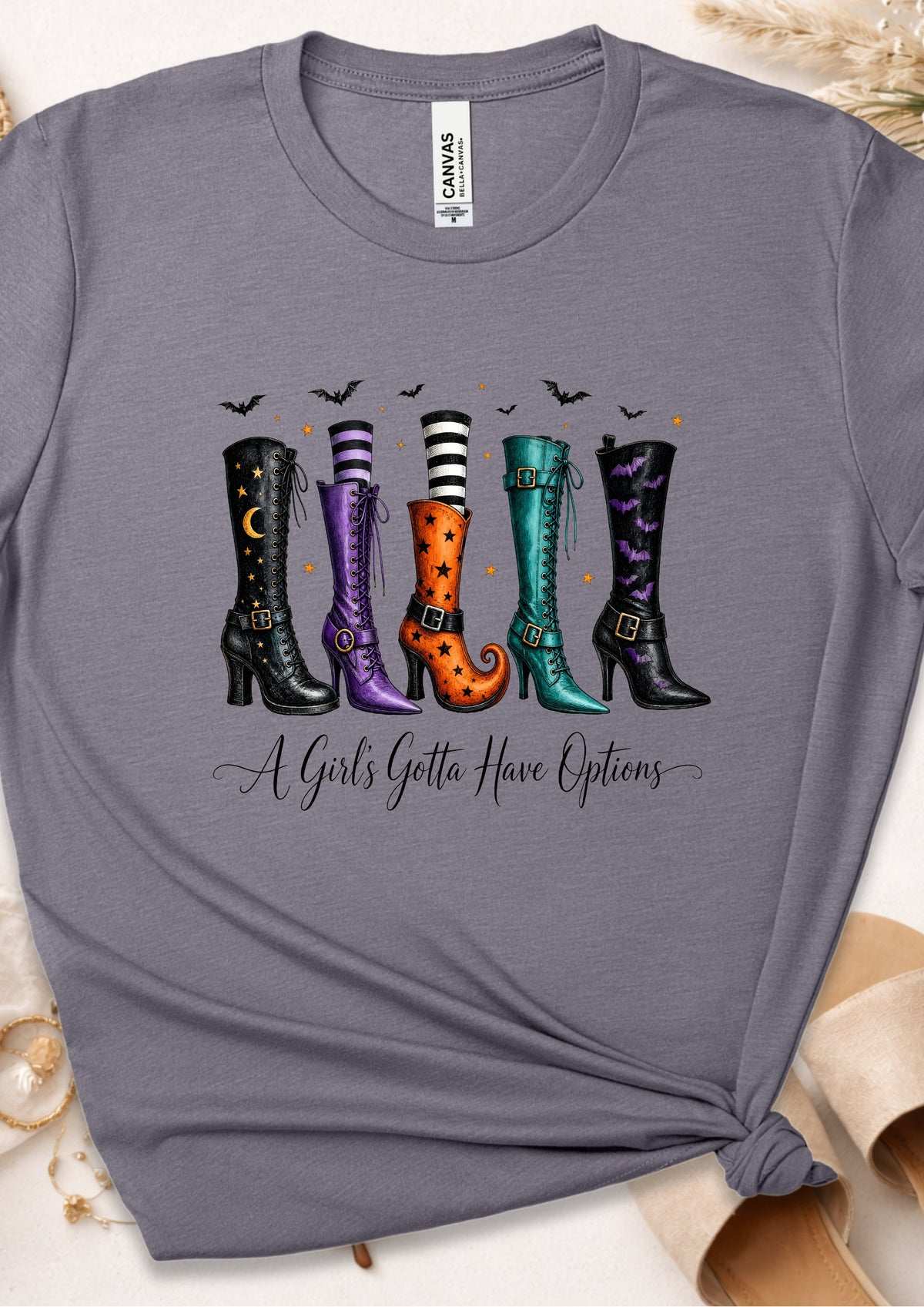 A heather gray graphic t-shirt featuring five colorful witch boot designs with the text 'A Girl's Gotta Have Options' underneath. The t-shirt is styled flat with a knot at the bottom hem, surrounded by neutral props.