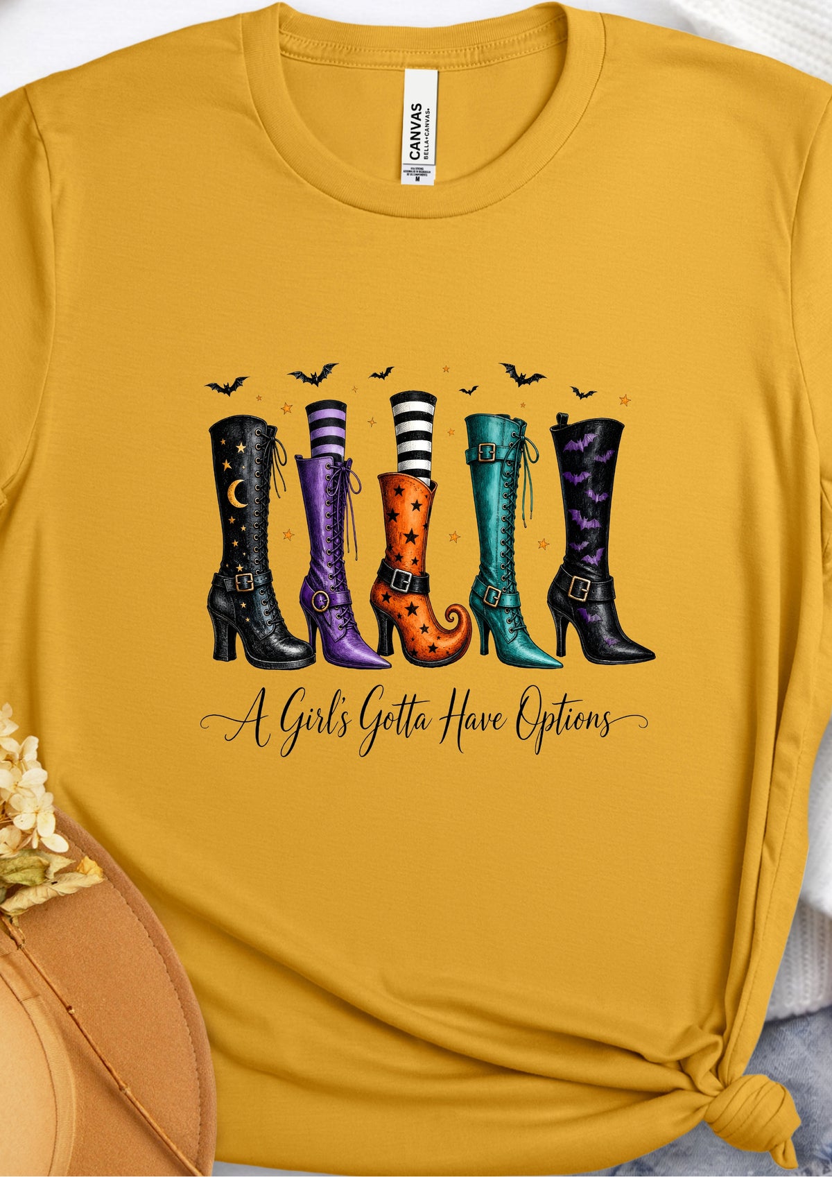 A mustard yellow graphic t-shirt featuring a colorful illustration of five witch boots in various patterns and colors, with bats and the text 'A Girl's Gotta Have Options' written below. A Gildan brand tag is visible inside the collar.