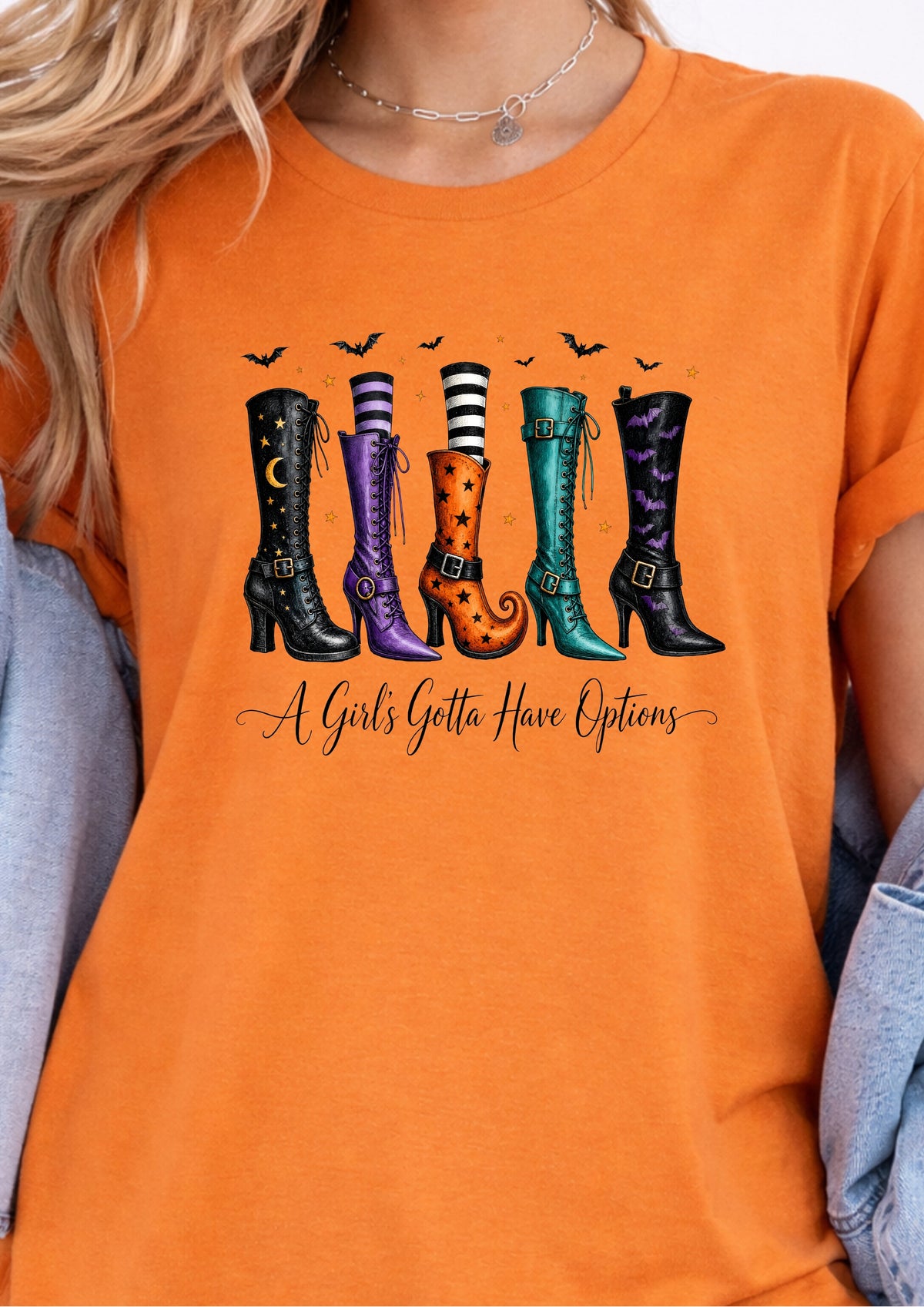 A woman wearing a bright orange graphic t-shirt. The shirt features a colorful illustration of six different witch-style high-heeled boots in black, purple, orange, and teal, with small black bats flying above and the phrase 'A Girls Getta Have Options' written in black script below.