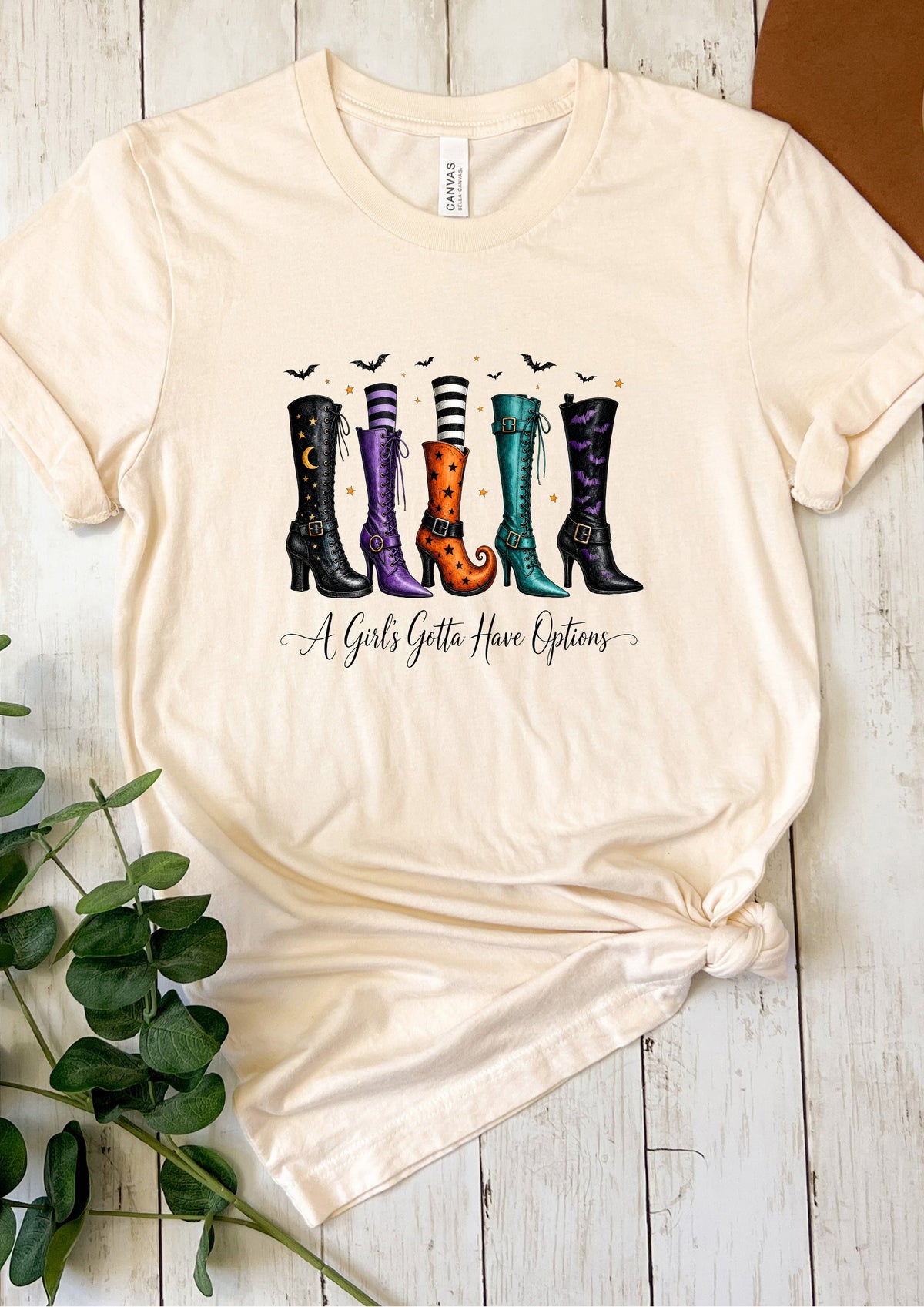 A cream-colored short-sleeve t-shirt with a graphic of four colorful witch boots and the text 'A Girls Gotta Have Options' underneath, styled on a white wooden background with a plant sprig.
