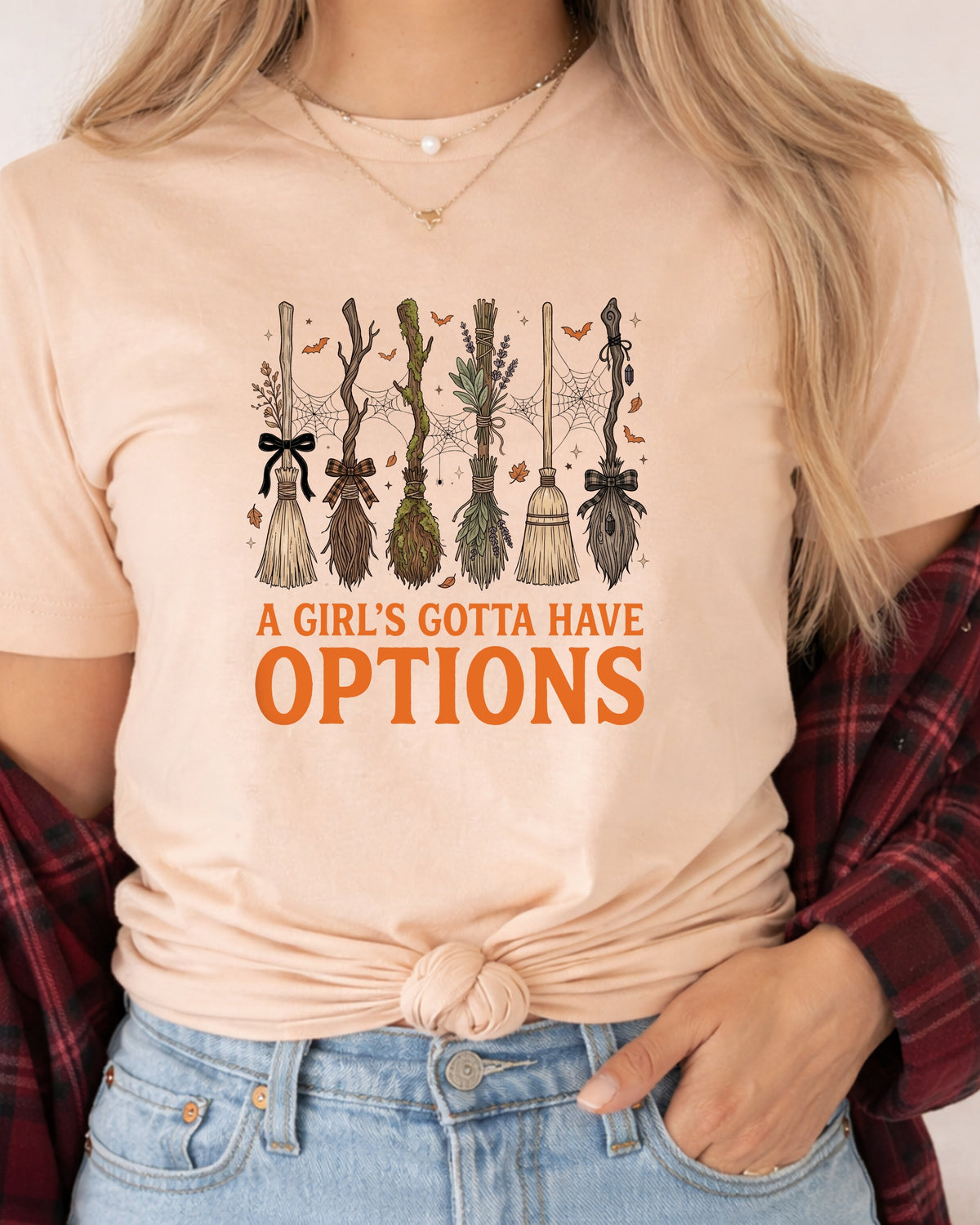 A woman wearing a light peach t-shirt with a graphic of five broomsticks and the text 'A GIRL'S GOTTA HAVE OPTIONS' in orange, styled with a red plaid shirt and blue jeans.