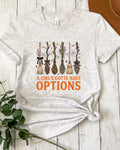 A light heather gray graphic t-shirt laid flat on a white wood plank background, styled with a green plant sprig on the left and a brown leather object in the top right corner. The t-shirt features a graphic of five broomsticks with autumn leaves and the text 'A GIRL'S GOTTA HAVE OPTIONS' in orange.
