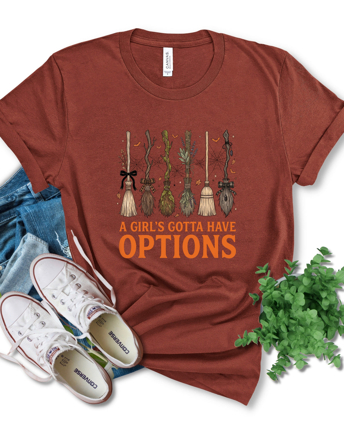 A rust-brown short-sleeve graphic T-shirt laid flat with jeans, white sneakers, and green leaves around it. The shirt has a Halloween-themed broom graphic and the text 'A GIRL'S GOTTA HAVE OPTIONS' printed on the front.