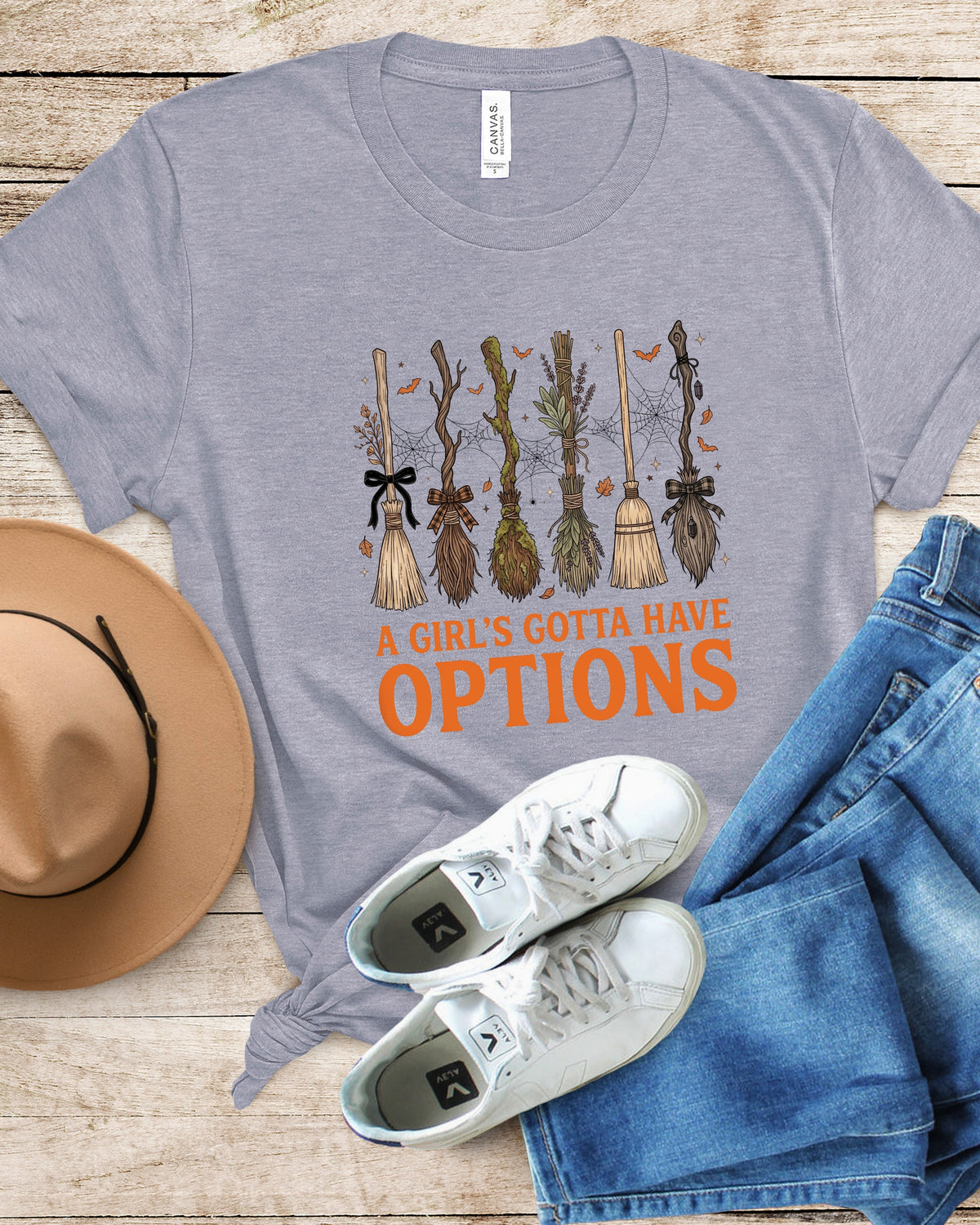 A heather gray graphic t-shirt featuring a Halloween-themed design with broomsticks and the text 'A GIRL'S GOTTA HAVE OPTIONS' in orange. The shirt is styled in a flat lay on a light wood background, accompanied by a tan wide-brim hat, blue jeans, and white sneakers.