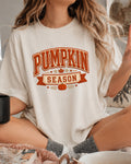 A woman wearing a cream-colored oversized t-shirt with a 'Pumpkin Season' graphic in orange and brown, sitting in a cozy indoor setting.