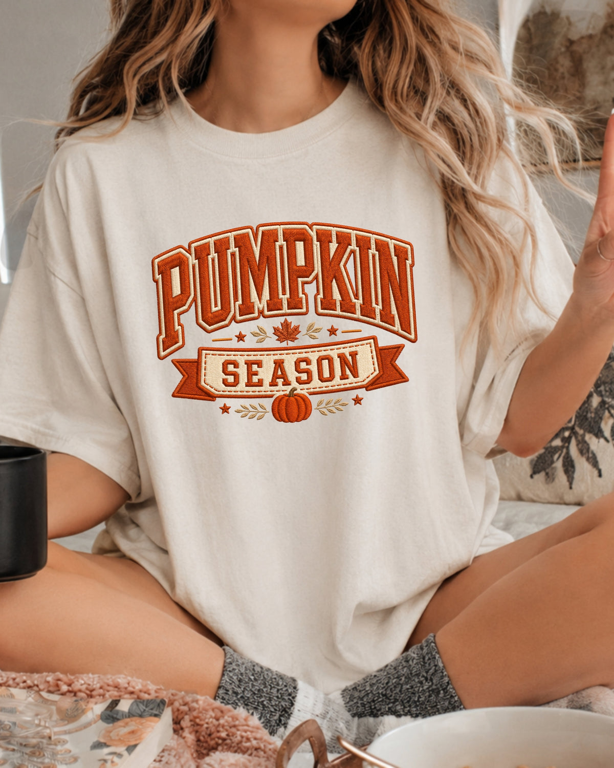 A woman wearing a cream-colored oversized t-shirt with a 'Pumpkin Season' graphic in orange and brown, sitting in a cozy indoor setting.