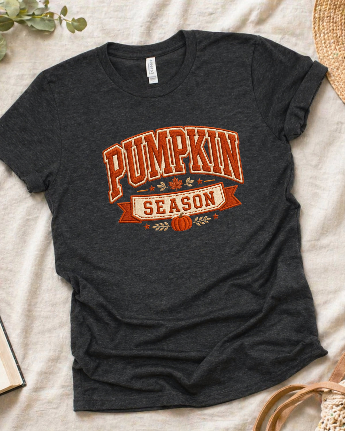 A dark heather charcoal t-shirt with a 'Pumpkin Season' graphic in orange and cream, laid flat on a beige fabric background with seasonal props.