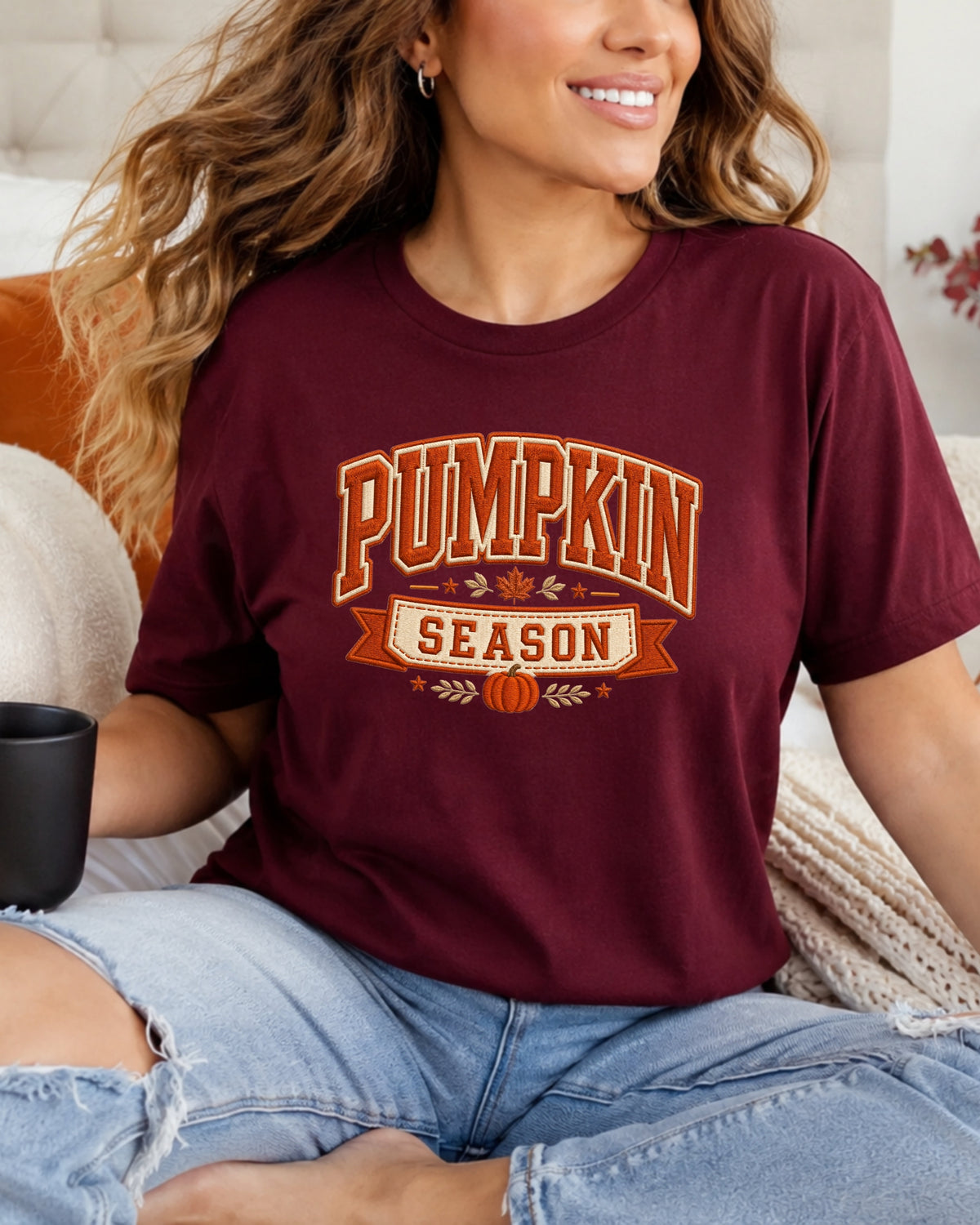 A woman wearing a maroon short-sleeve graphic T-shirt with 'PUMPKIN SEASON' printed in orange and white, paired with light-wash ripped jeans, sitting on a couch.